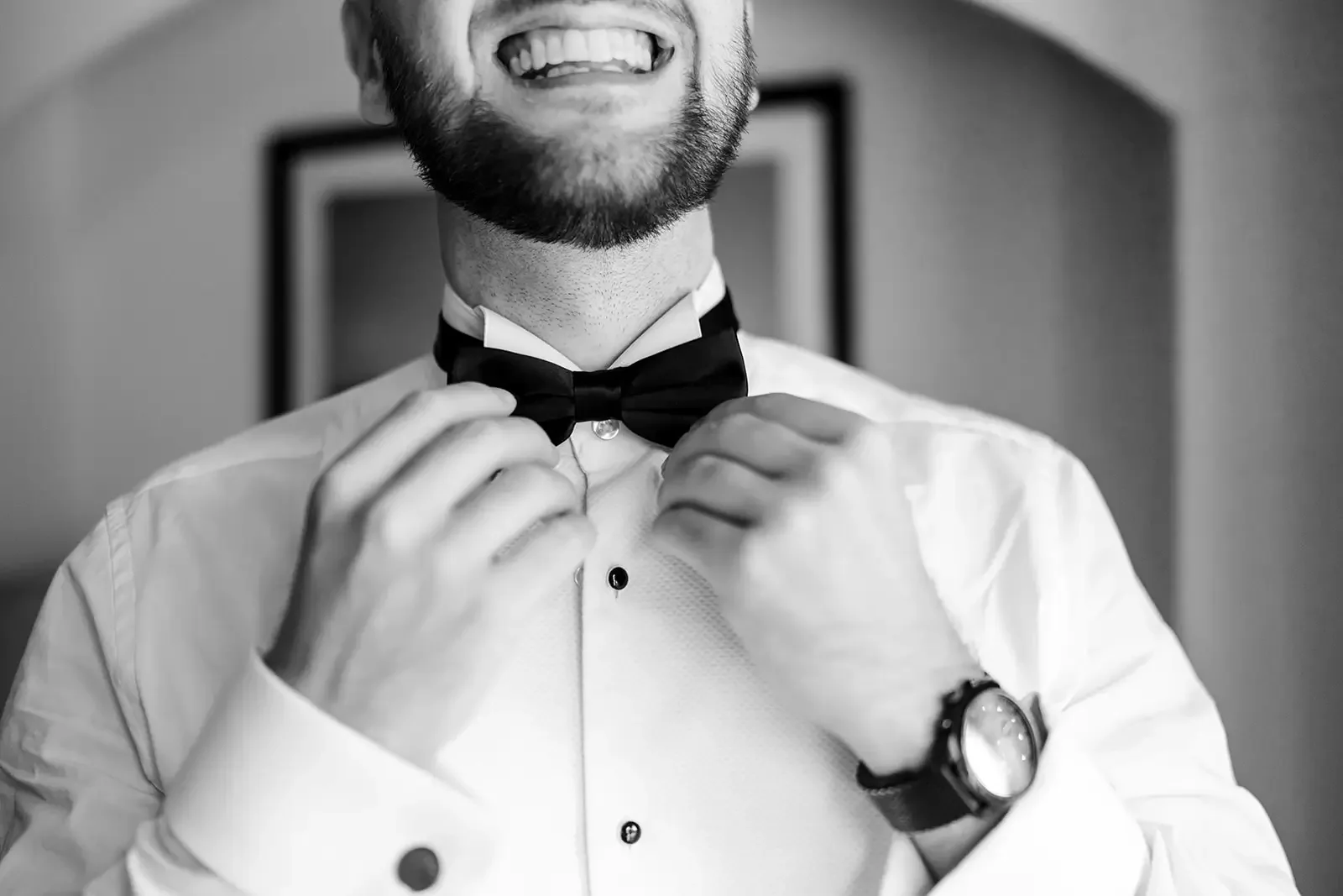 A man adjusting a black bow tie, wearing a white dress shirt and a wristwatch, smiling with his mouth open.