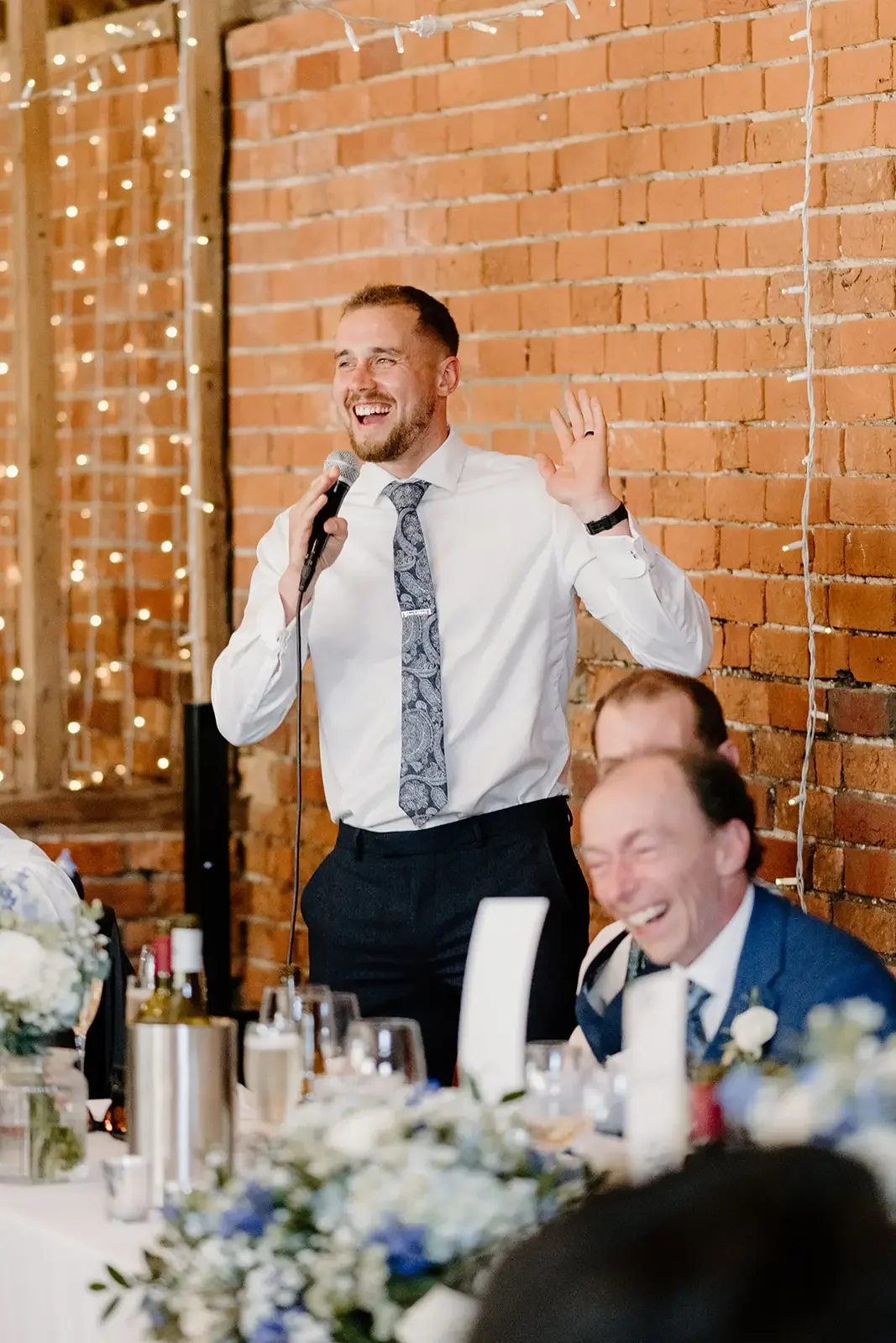 A man in a white shirt and patterned tie is giving a speech with a microphone, smiling and waving, at a celebration or wedding reception with a brick wall and string lights in the background. Others are seated at a table decorated with flowers and ca