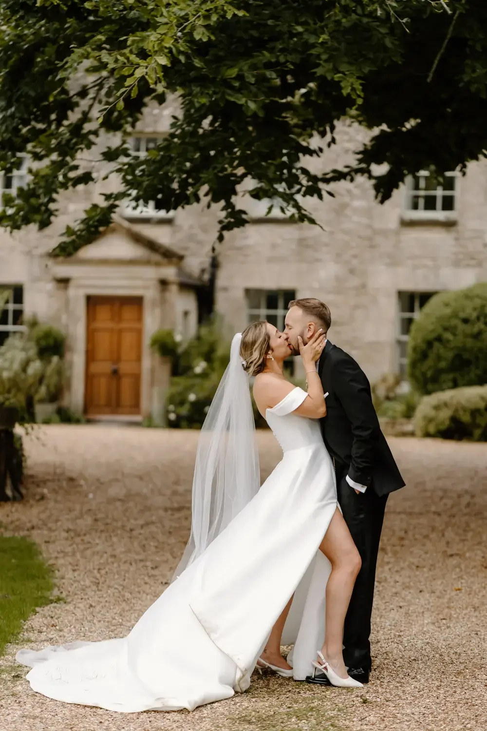 A newlywed couple sharing a kiss outdoors in front of a stone house, with the bride wearing a white off-shoulder wedding gown and veil, and the groom in a black tuxedo.