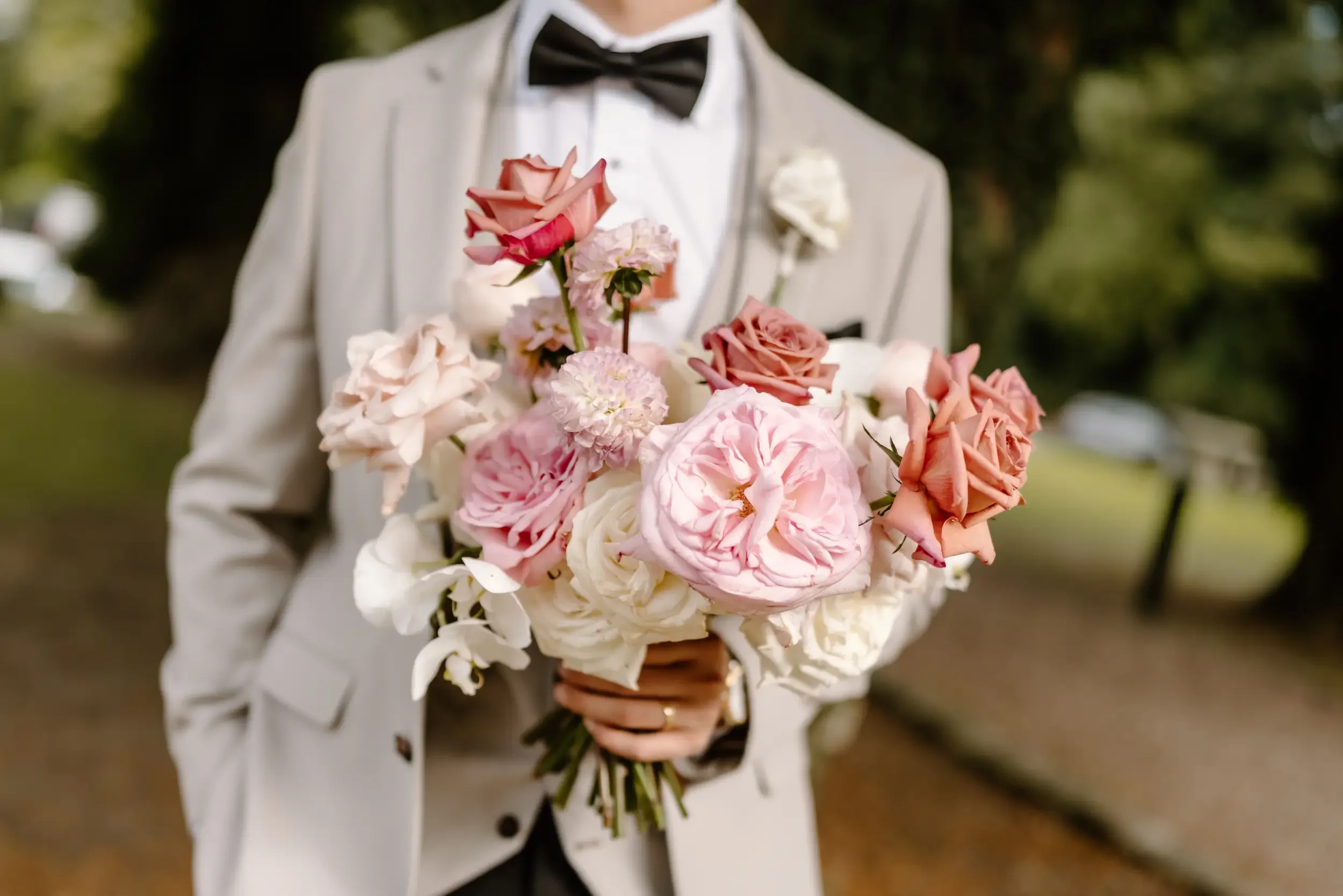 Person in a light-colored suit holding a bouquet of pink, white, and peach roses and other flowers outdoors.
