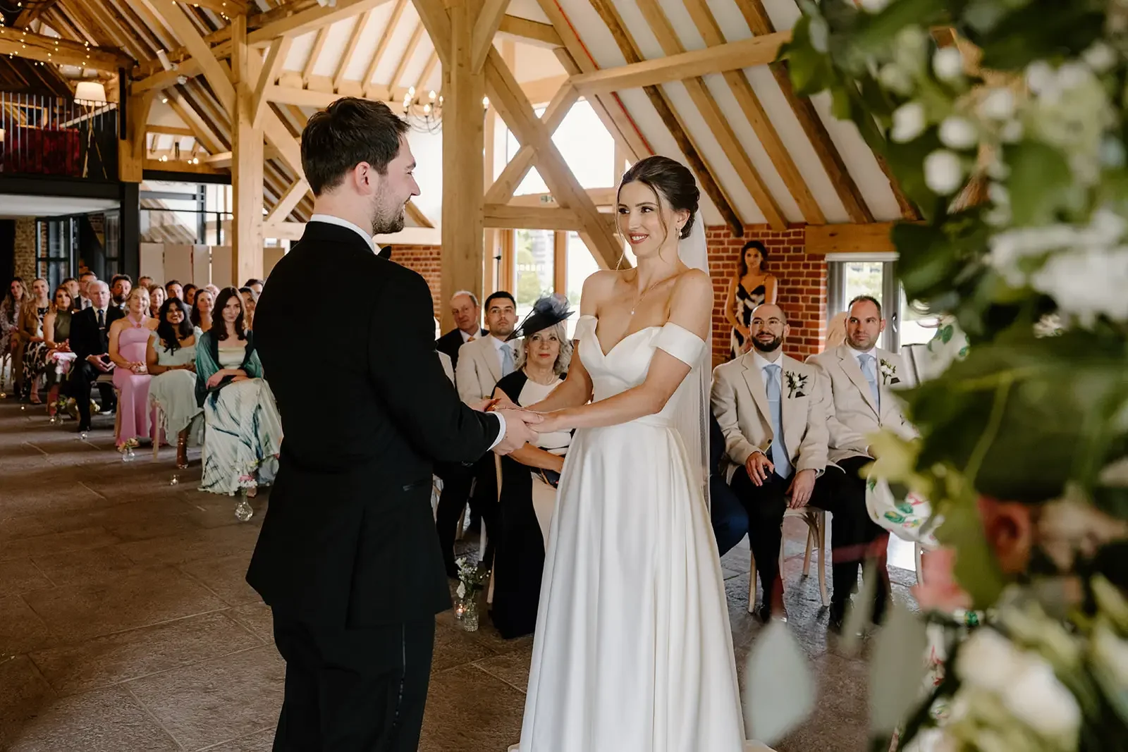 A wedding ceremony with a bride and groom holding hands and exchanging vows in a rustic indoor venue, with guests seated and watching.