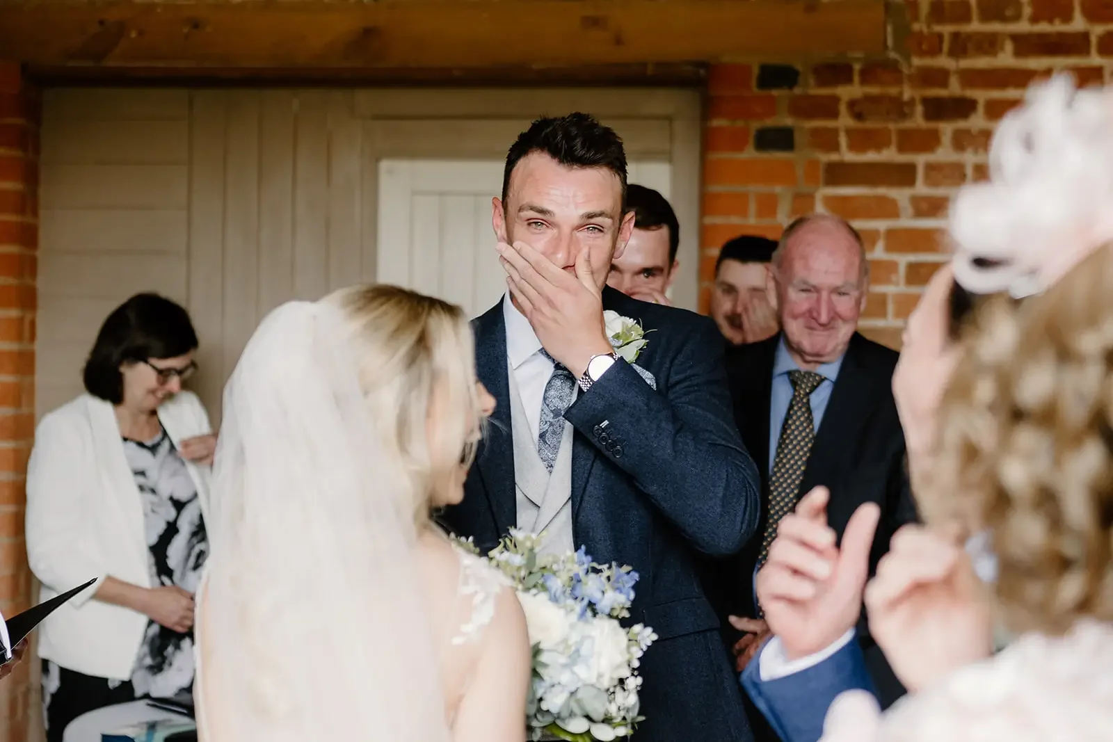 A groom in a dark suit covering his mouth with his hand, smiling, during a wedding ceremony. The bride is partially visible holding a bouquet. Several well-dressed guests stand in the background in a rustic setting with exposed brick walls.