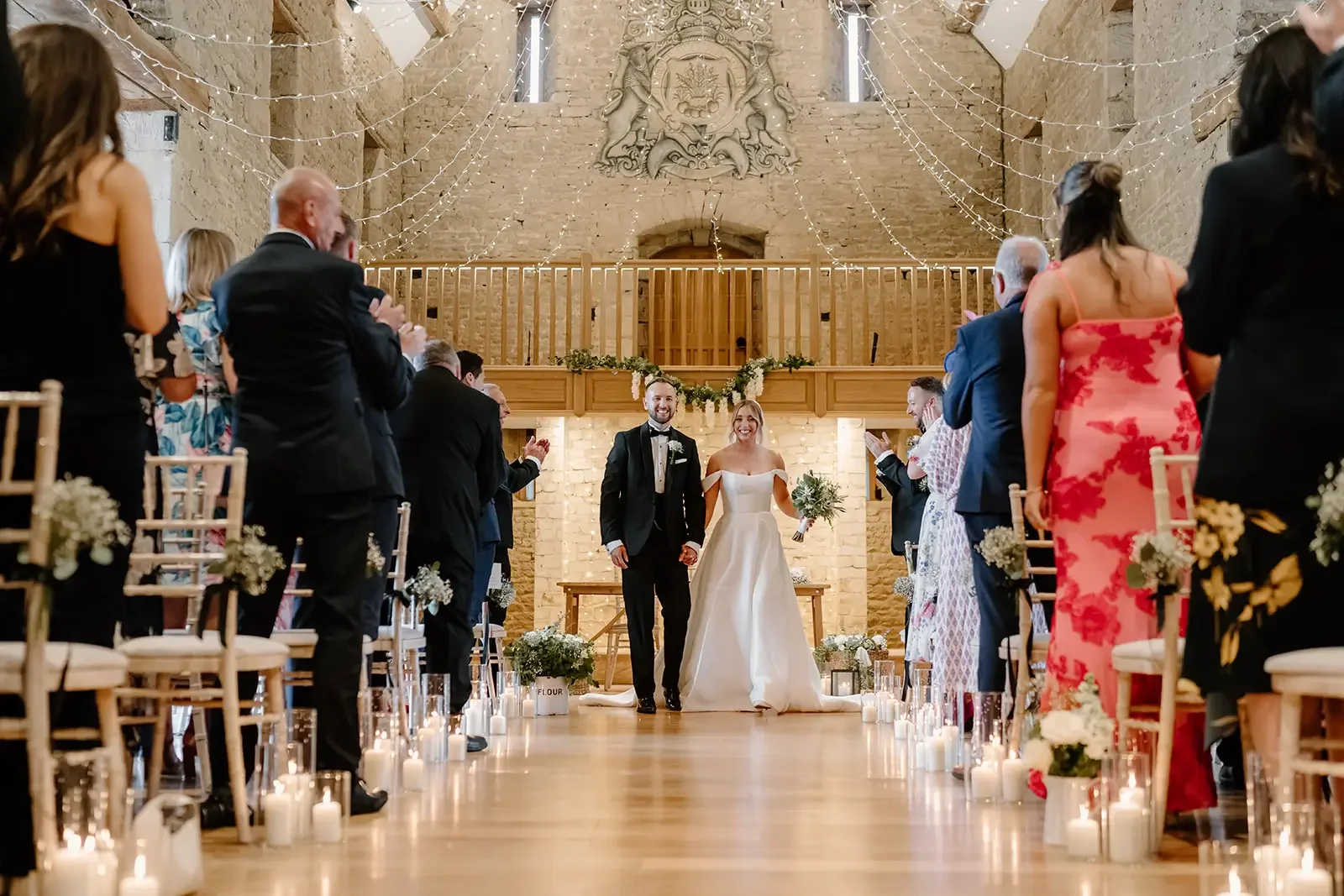 A bride and groom walking down the aisle together in a wedding ceremony inside a rustic brick venue, surrounded by seated guests on either side, with candles and floral decorations.