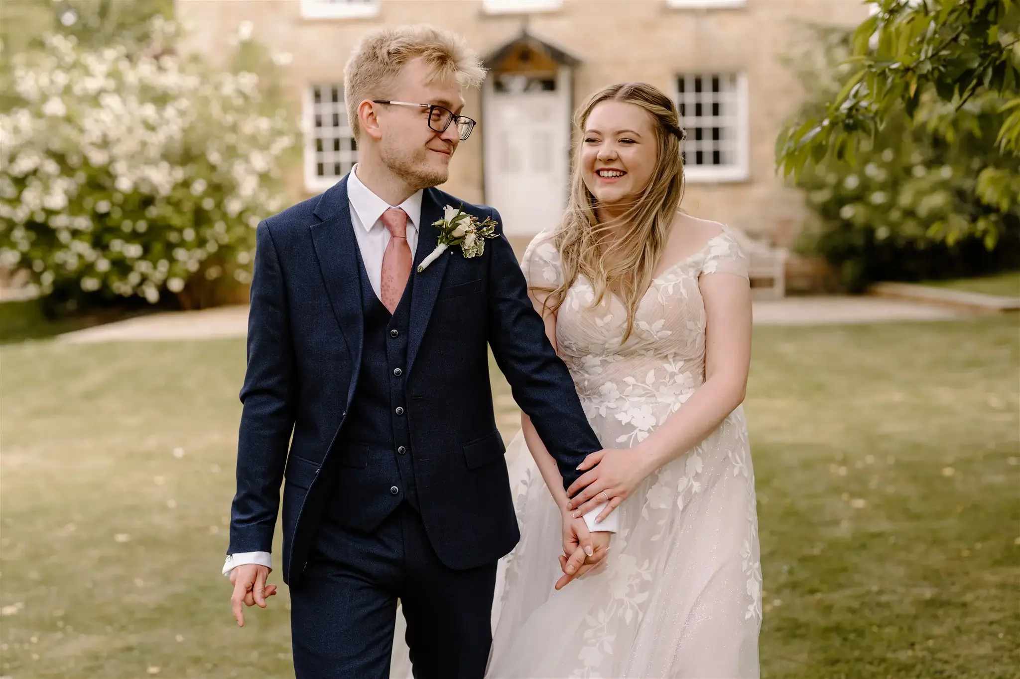Bride and groom walking hand in hand outdoors, smiling and holding hands, with a house and greenery in the background.