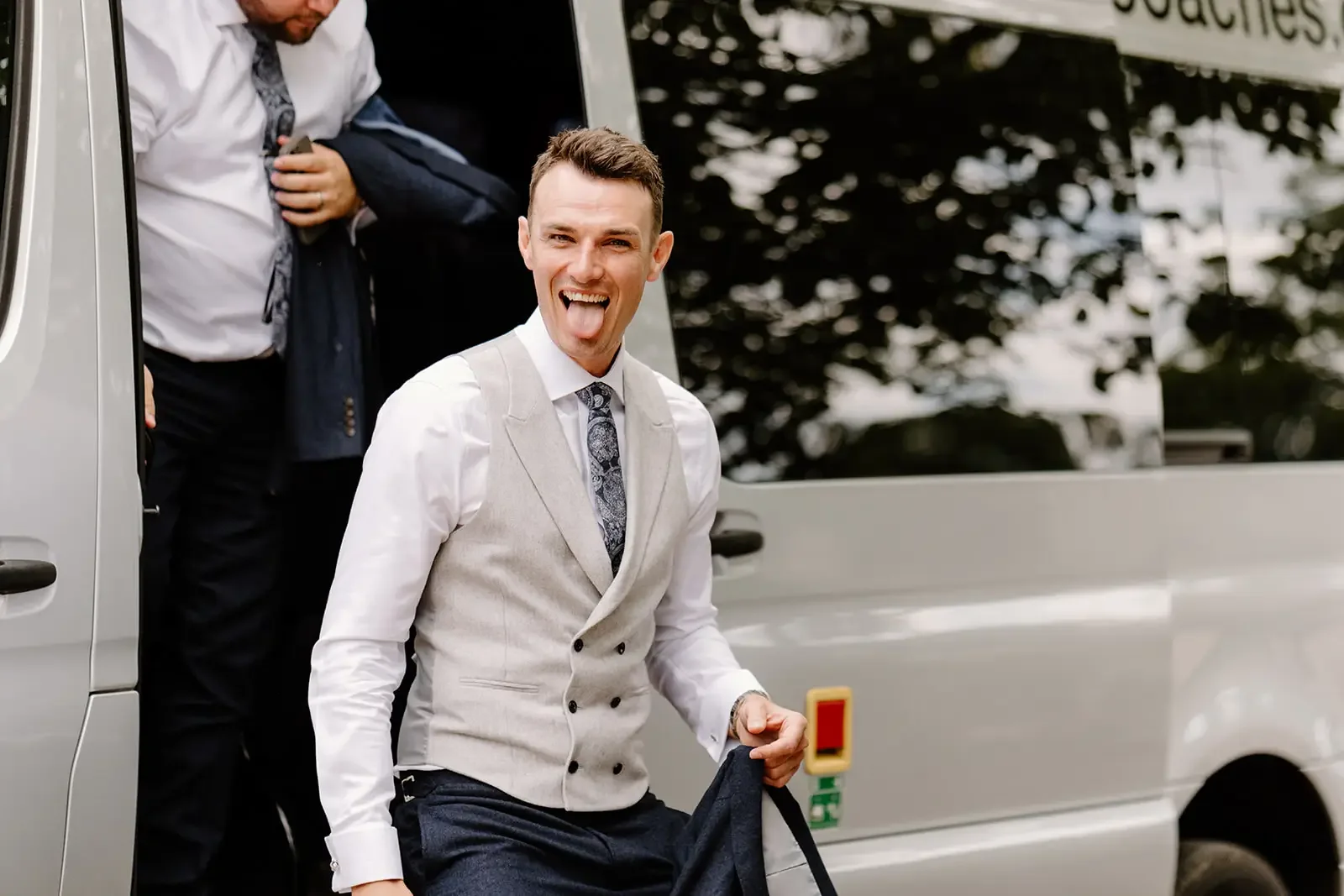 A smiling man with short brown hair is standing outside a vehicle, sticking out his tongue, wearing a white shirt, a light gray vest, and a patterned tie. He is holding a dark garment, and behind him, another man is disembarking from the vehicle, adj