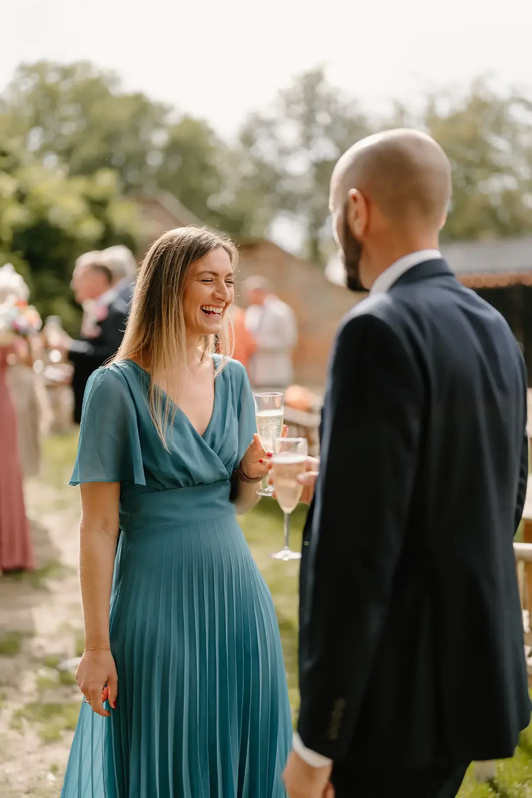 A woman in a blue dress holding a glass of champagne, smiling and laughing while talking to a man in a dark suit at an outdoor wedding reception.