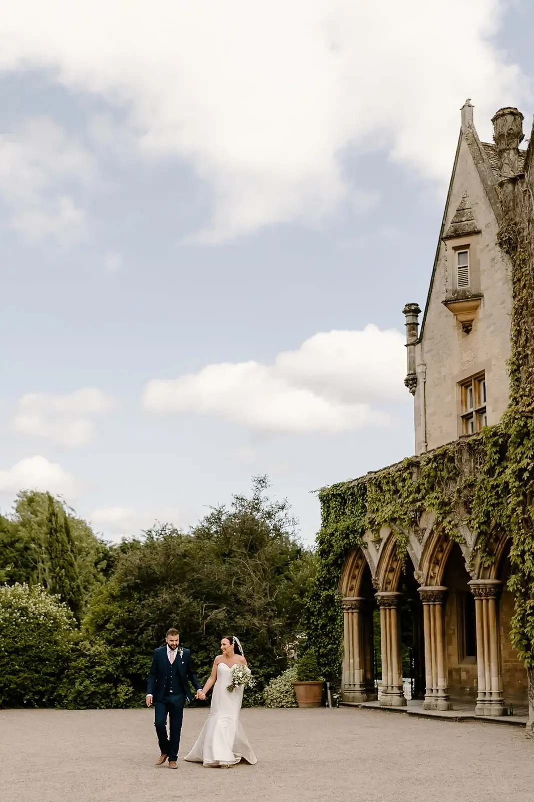 A bride and groom walking hand in hand outside a historic building with ivy-covered arches on a cloudy day.
