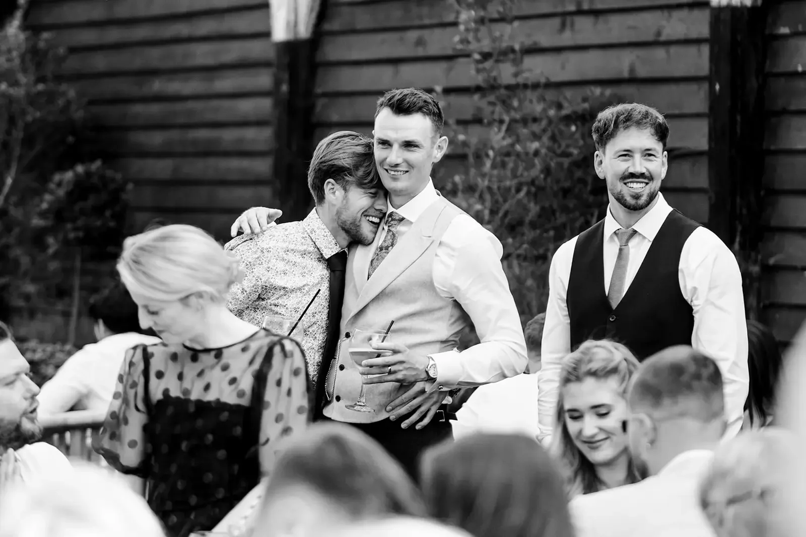 Group of people at a celebration, with three men standing and smiling, one holding a drink, in an outdoor setting.