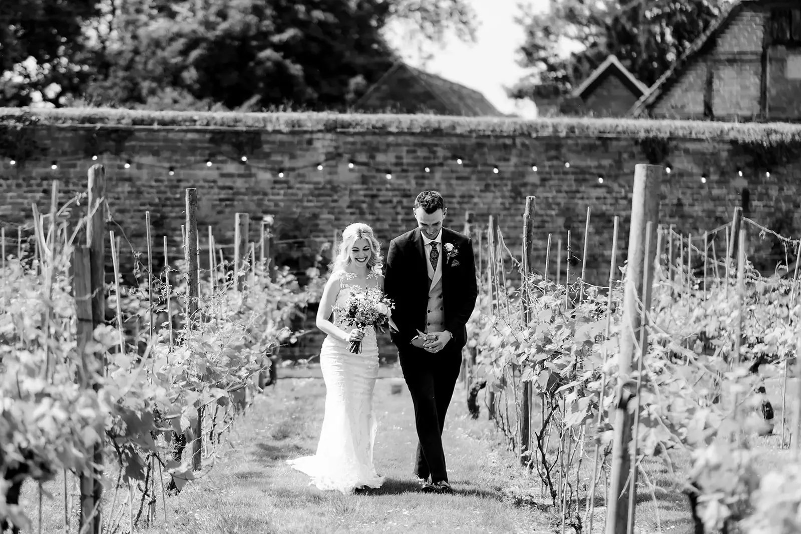 A bride and groom walking together in a vineyard, smiling, with the bride holding a bouquet, black and white photograph.