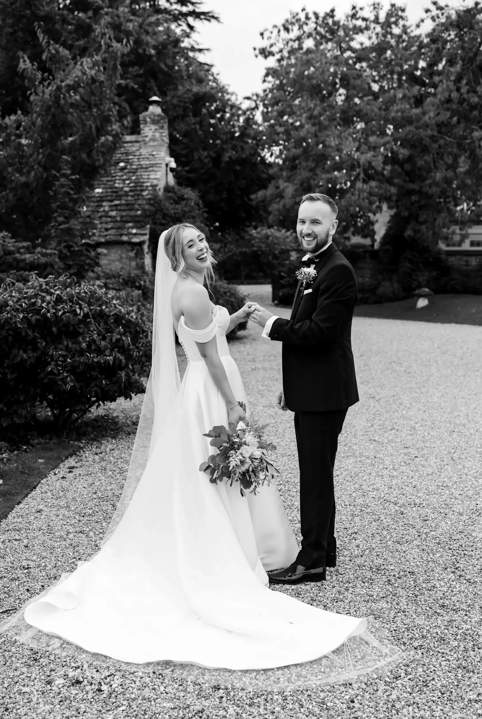 Black and white photo of a bride and groom holding hands and smiling outdoors, with trees and a stone structure in the background.
