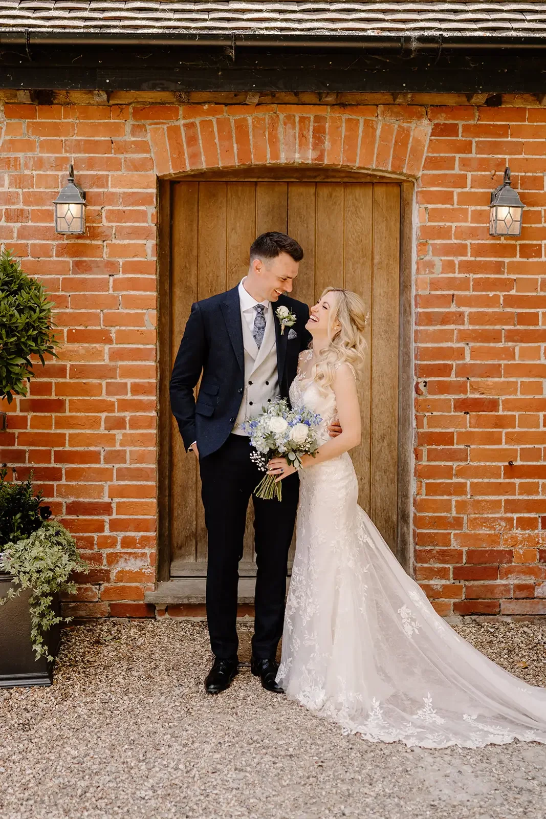 A bride and groom on their wedding day standing in front of a wooden door, smiling and looking at each other, with the bride holding a bouquet of white and blue flowers, against a brick wall with outdoor lanterns.