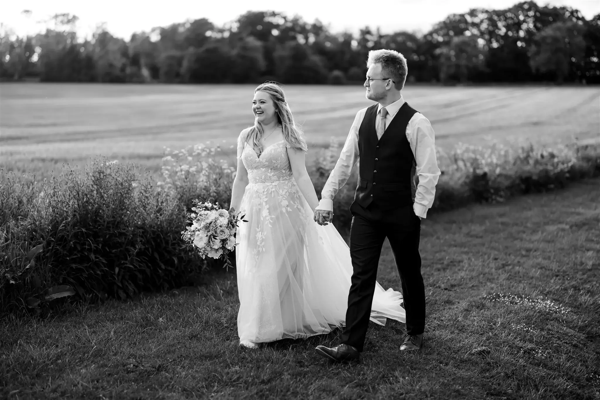 A bride and groom holding hands and walking across a grassy field during their wedding in black and white.
