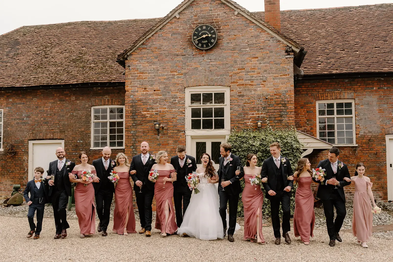 Group of people dressed in wedding attire, walking arm-in-arm in front of a rustic brick building with a clock on the facade and windows.