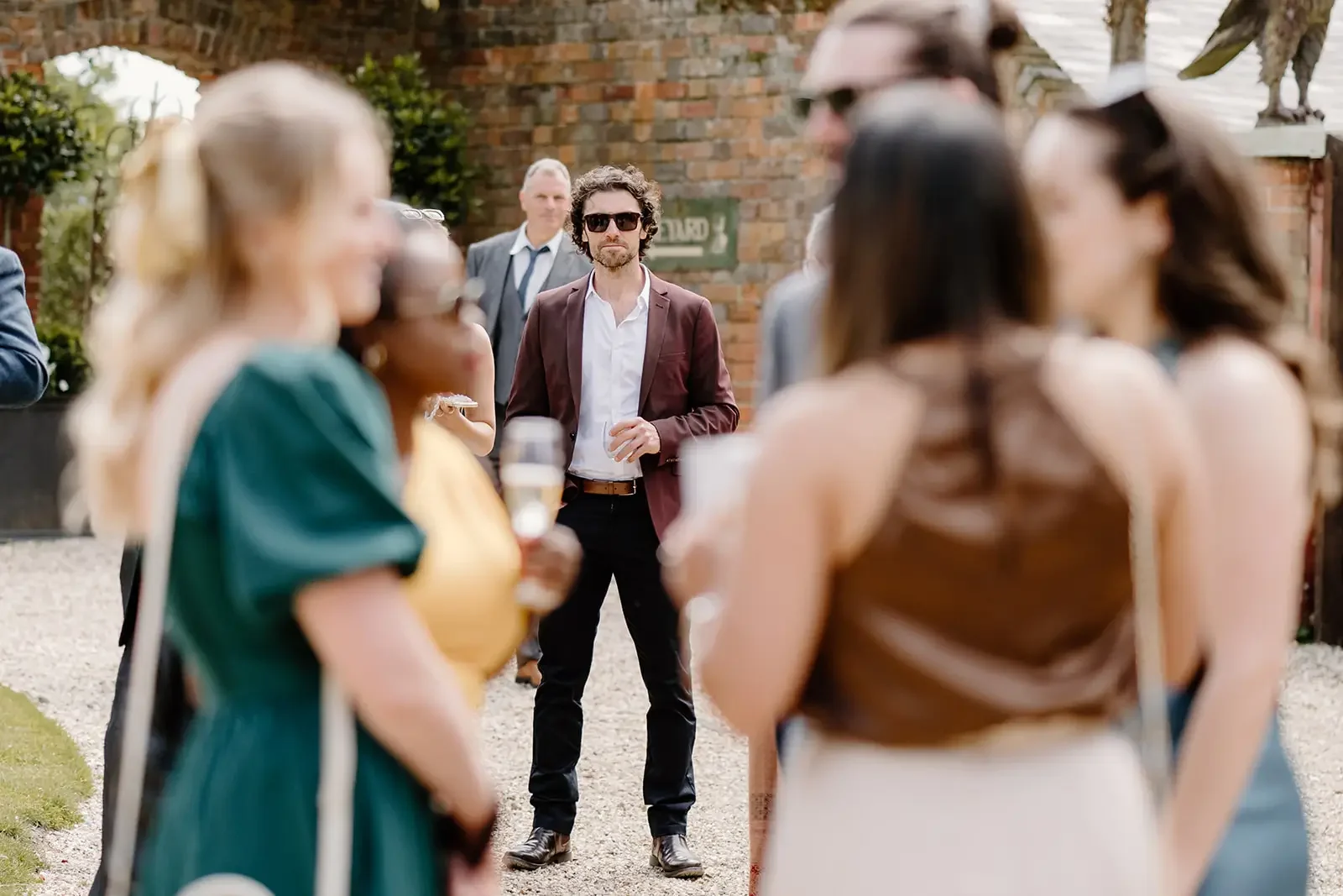 A man in sunglasses and a burgundy blazer stands among a group of women holding drinks at an outdoor social gathering, with a brick wall and greenery in the background.