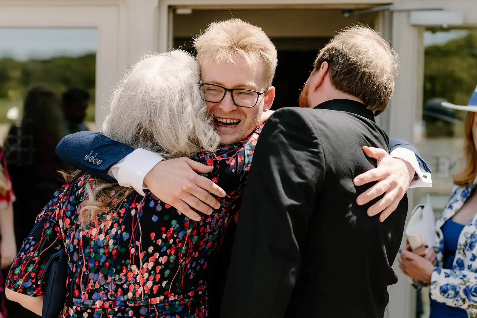 A young man with glasses is hugging two older women, one with grey hair and the other with darker hair, outside a building with glass windows, smiling happily.