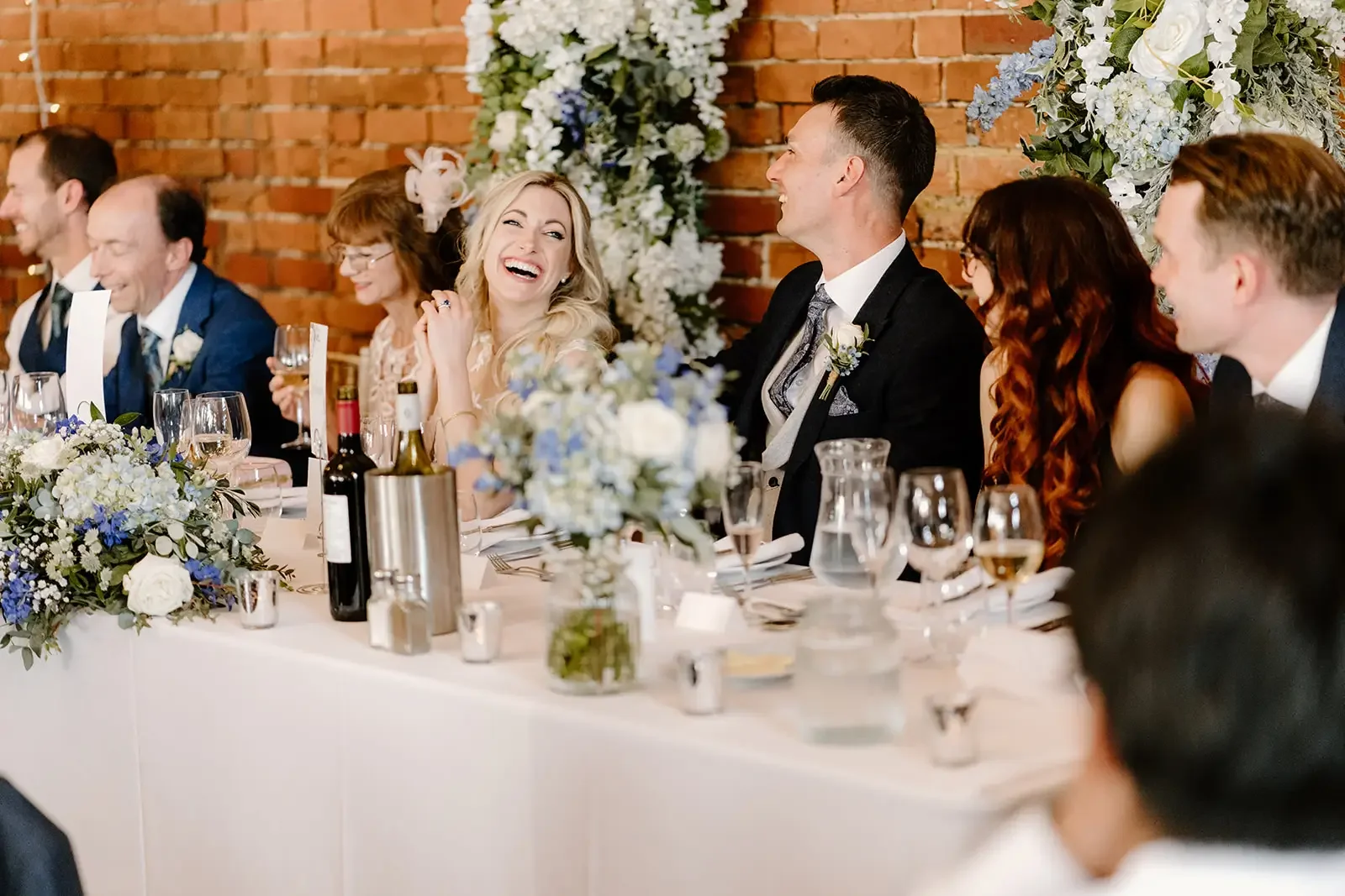 Wedding reception with guests sitting at a long table, smiling, and laughing, decorated with flowers and drinks, in front of a brick wall.
