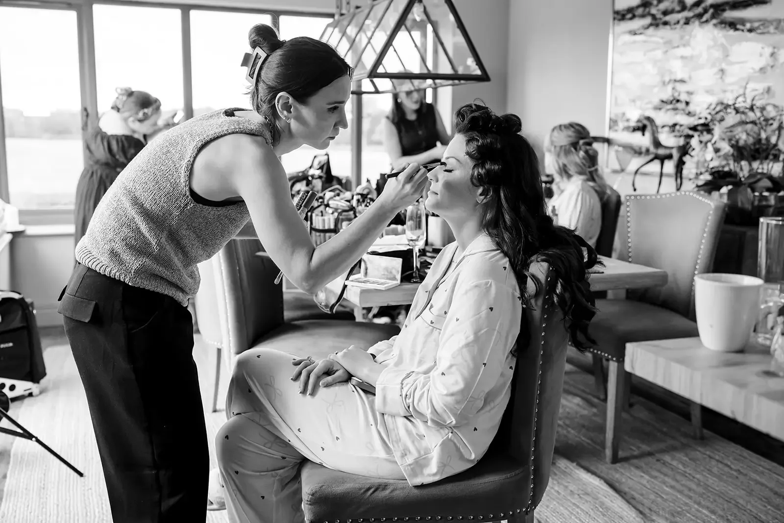 A woman is sitting in a chair with her eyes closed while a makeup artist applies makeup to her face in a room with large windows and other women preparing in the background.