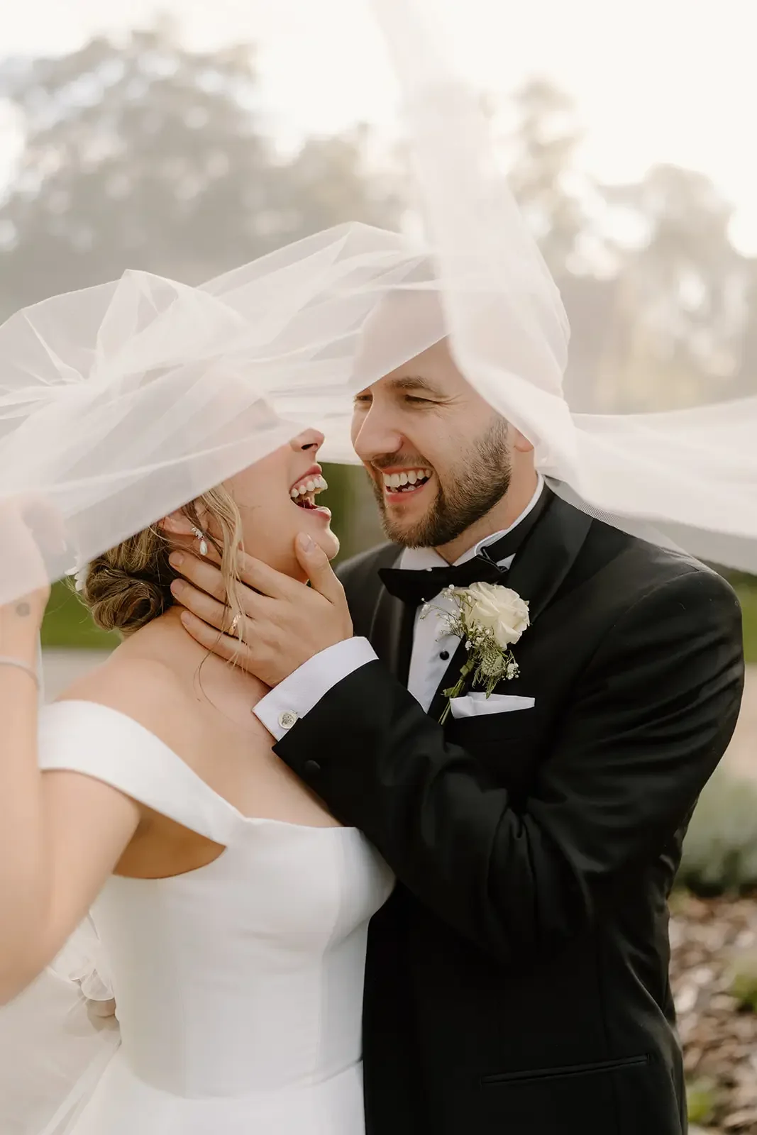A newlywed couple shares a joyful moment outdoors, with the bride in a white wedding dress and the groom in a black tuxedo, under a sheer fabric veil.