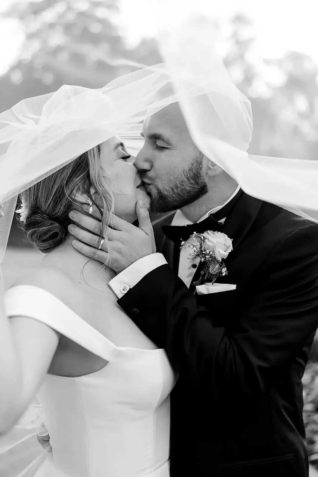 A bride and groom sharing a kiss, with the bride wearing a wedding dress and a veil, and the groom in a tuxedo with a boutonnière, outside with trees in the background.