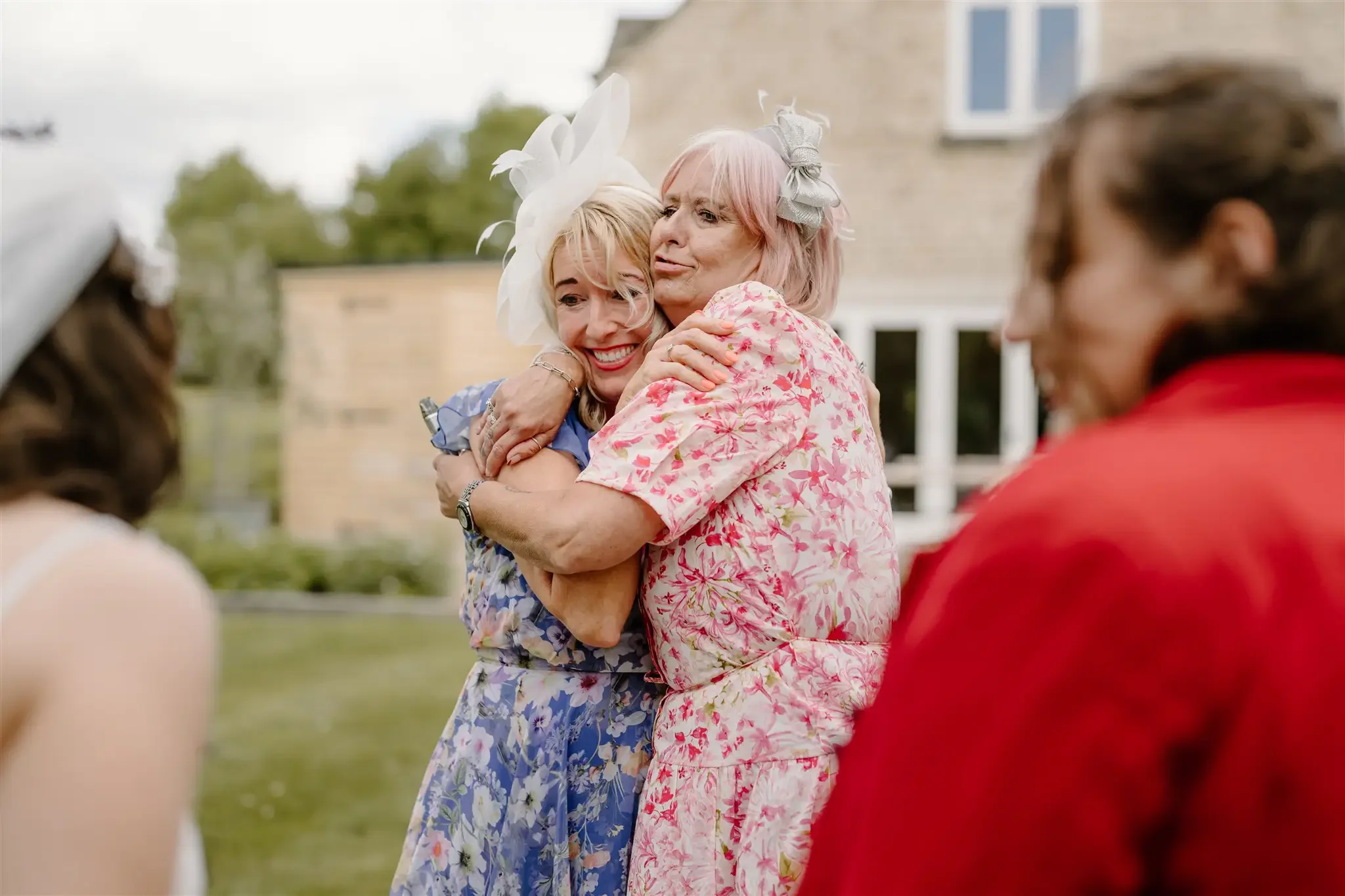 Two women wearing floral dresses and hats hugging each other while smiling, with two other women partially visible and blurred in the foreground. They are outdoors, in front of a house with a yard.