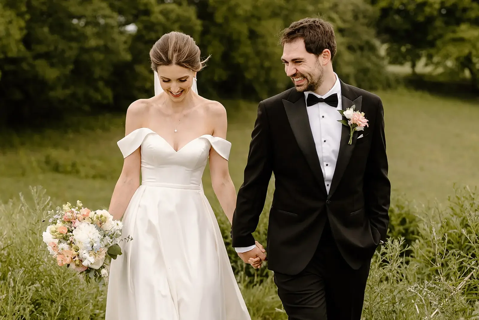 A bride and groom walking hand in hand outdoors during wedding, smiling, with the bride holding a bouquet of flowers and wearing a white wedding dress, and the groom in a black tuxedo with a boutonniere, surrounded by greenery.