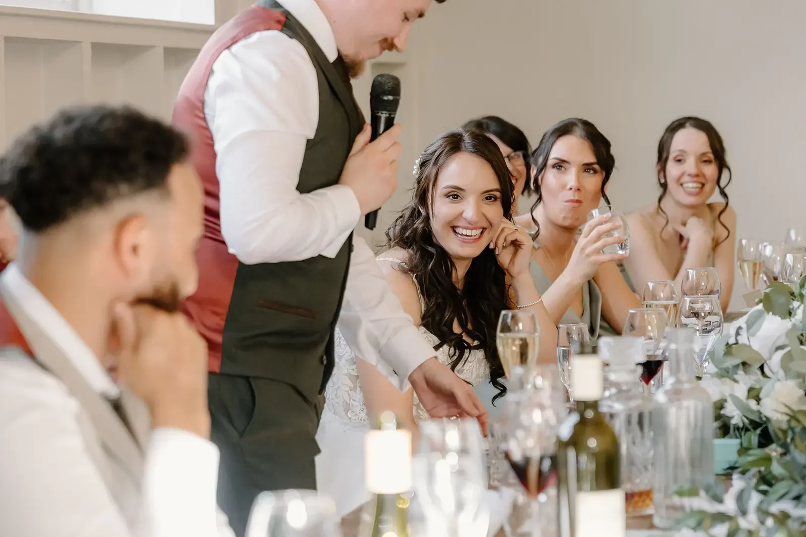 A man in a suit giving a speech at a wedding reception, with smiling women sitting at the table listening and enjoying.