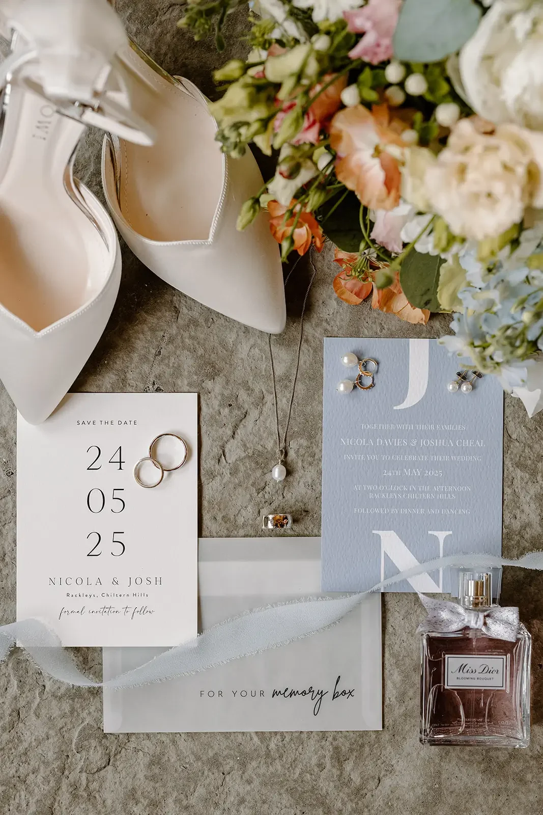 Wedding invitation, jewelry, floral bouquet, white high heels, Miss Dior perfume, and a memory box on a stone surface.