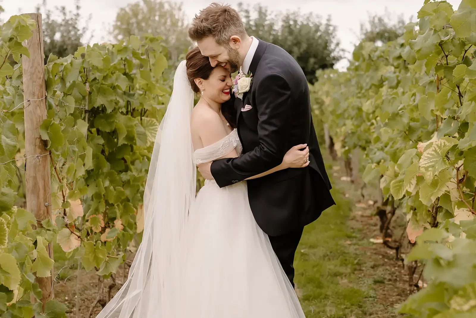 A bride and groom smiling and embracing in a vineyard. The bride is in a white wedding dress with a veil, and the groom is in a black tuxedo. They are surrounded by green grapevines.