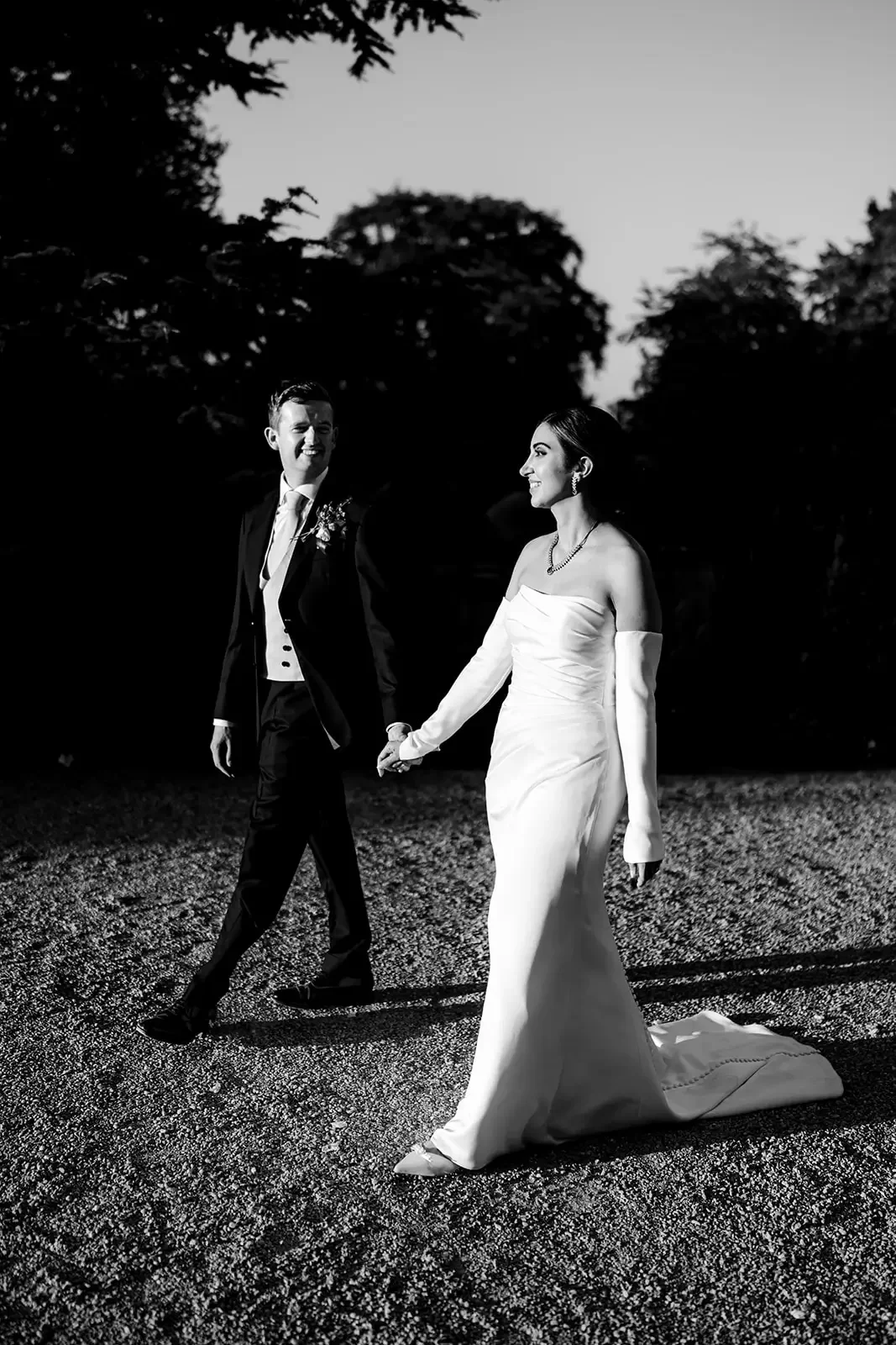 A black and white photo of a bride and groom walking hand in hand outdoors during dusk or dawn, with trees in the background.
