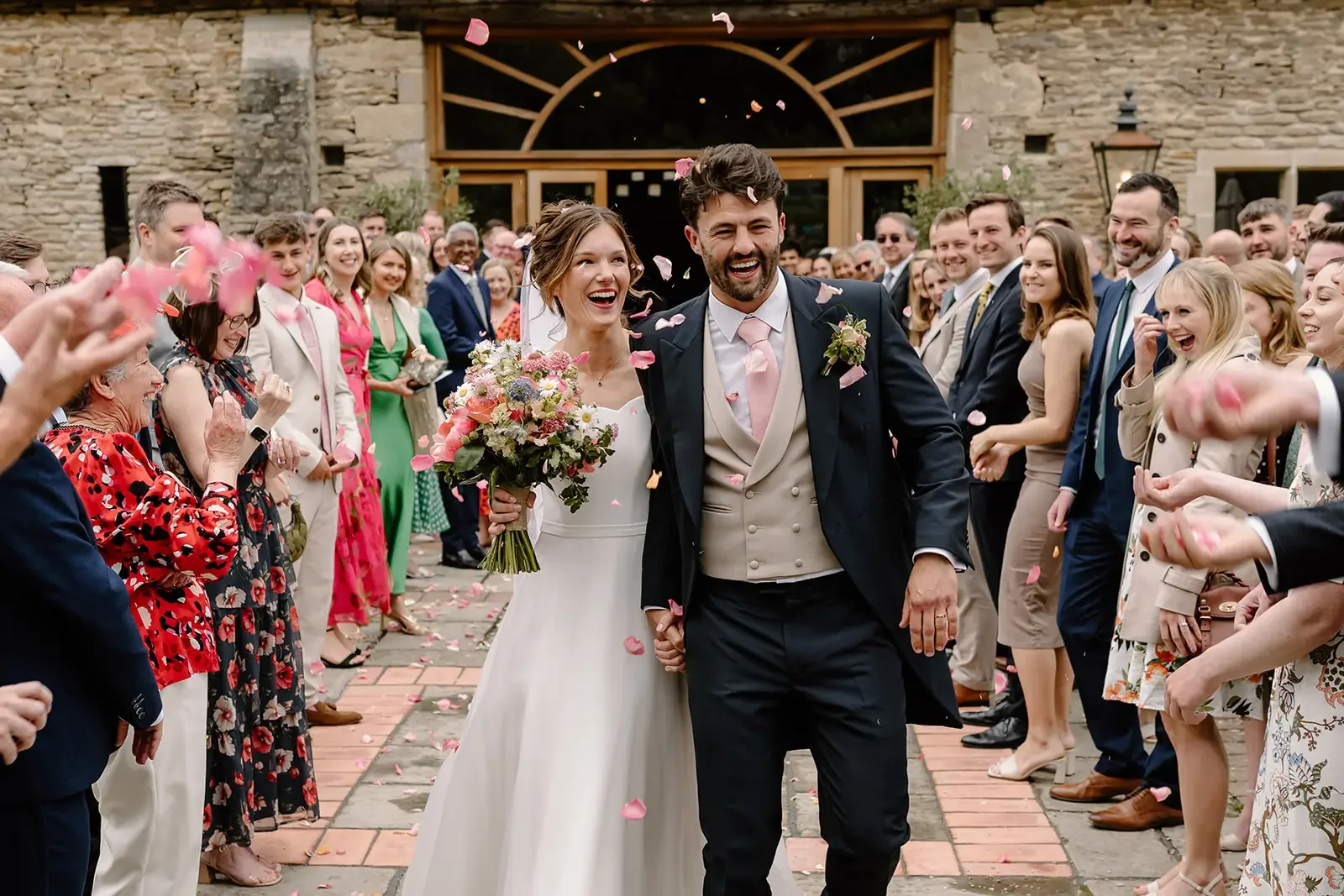 A bride and groom walking hand in hand after their wedding ceremony, surrounded by friends and family throwing flower petals, outside a rustic building.
