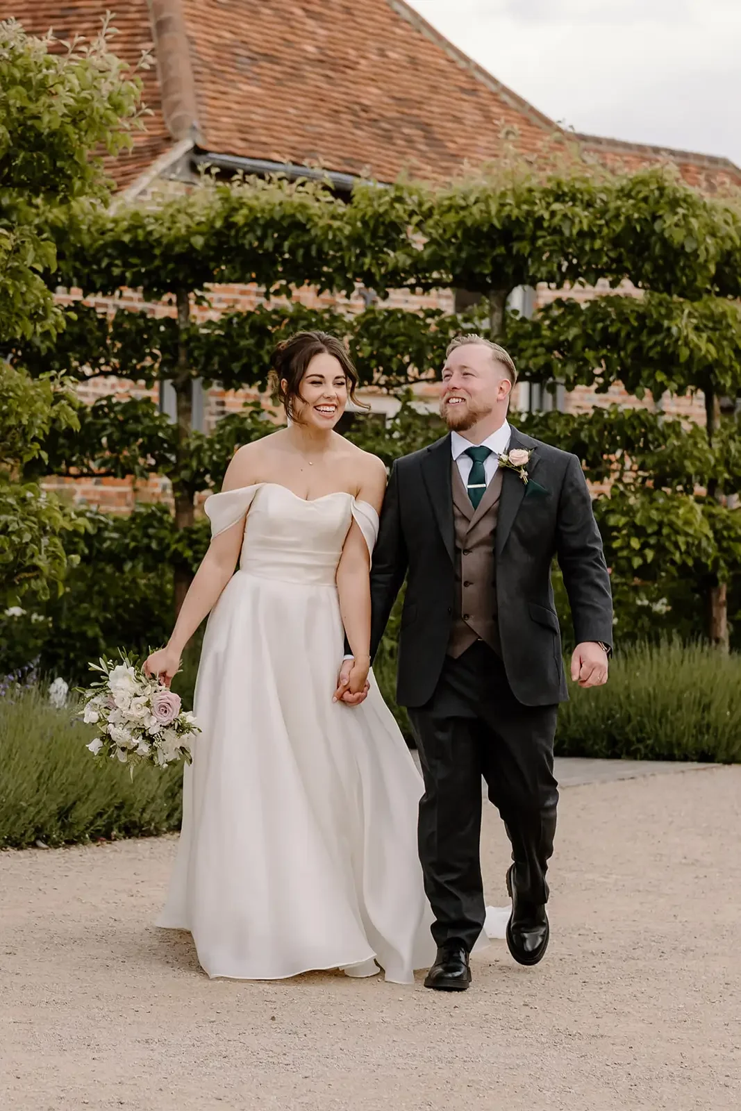 A bride and groom walking hand in hand outdoors, smiling and happy, with green trees and a brick building in the background.