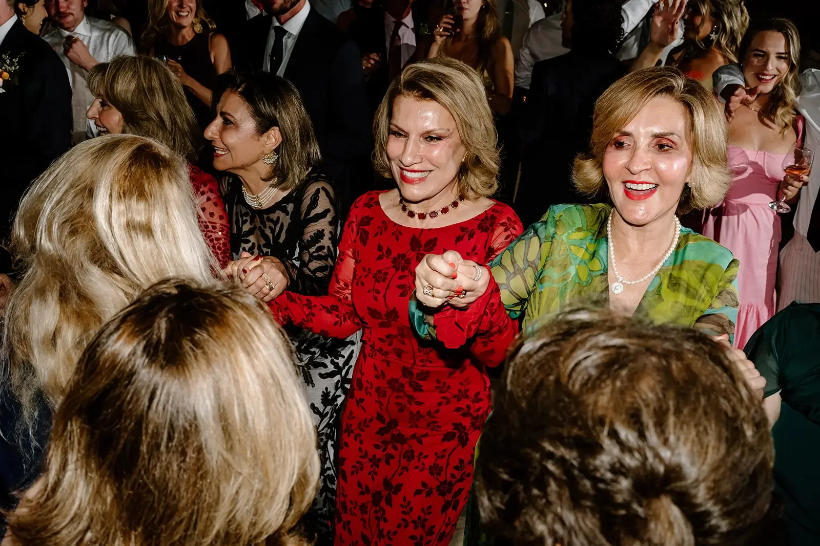 A group of women at a social gathering, with three women in the foreground smiling and holding hands.