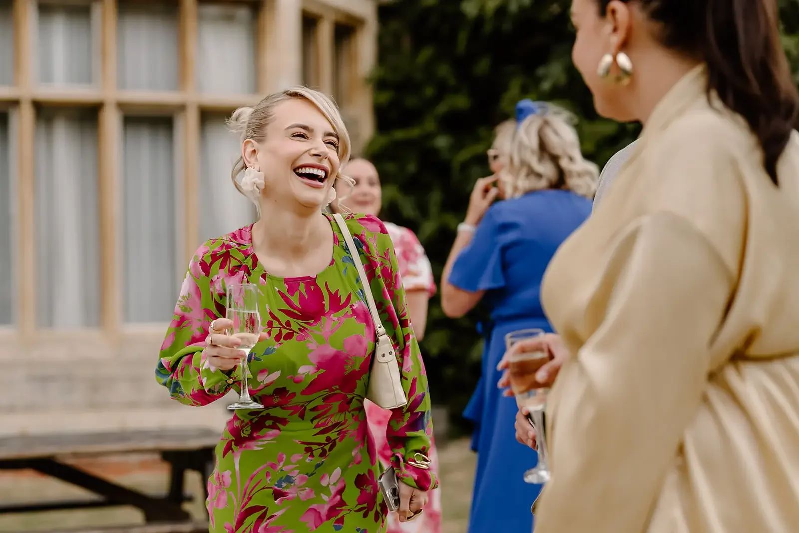 Group of women at an outdoor event, engaging in conversation and holding drinks, with a woman in a pink and green floral dress smiling.