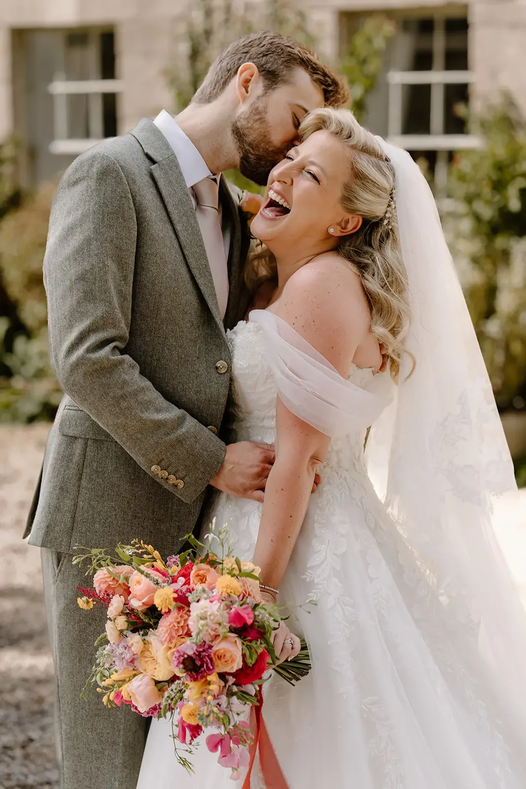 A bride and groom share a joyful moment outdoors, with the groom kissing the bride's forehead. The bride is smiling and holding a colorful bouquet, while the groom is wearing a gray suit. The bride is in a white wedding dress and veil.