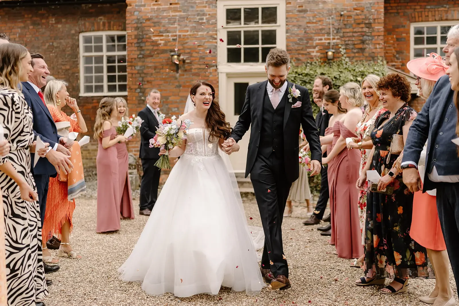 Bride and groom walking hand-in-hand in front of wedding guests outside a brick building, surrounded by smiling friends and family, with flower petals in the air.