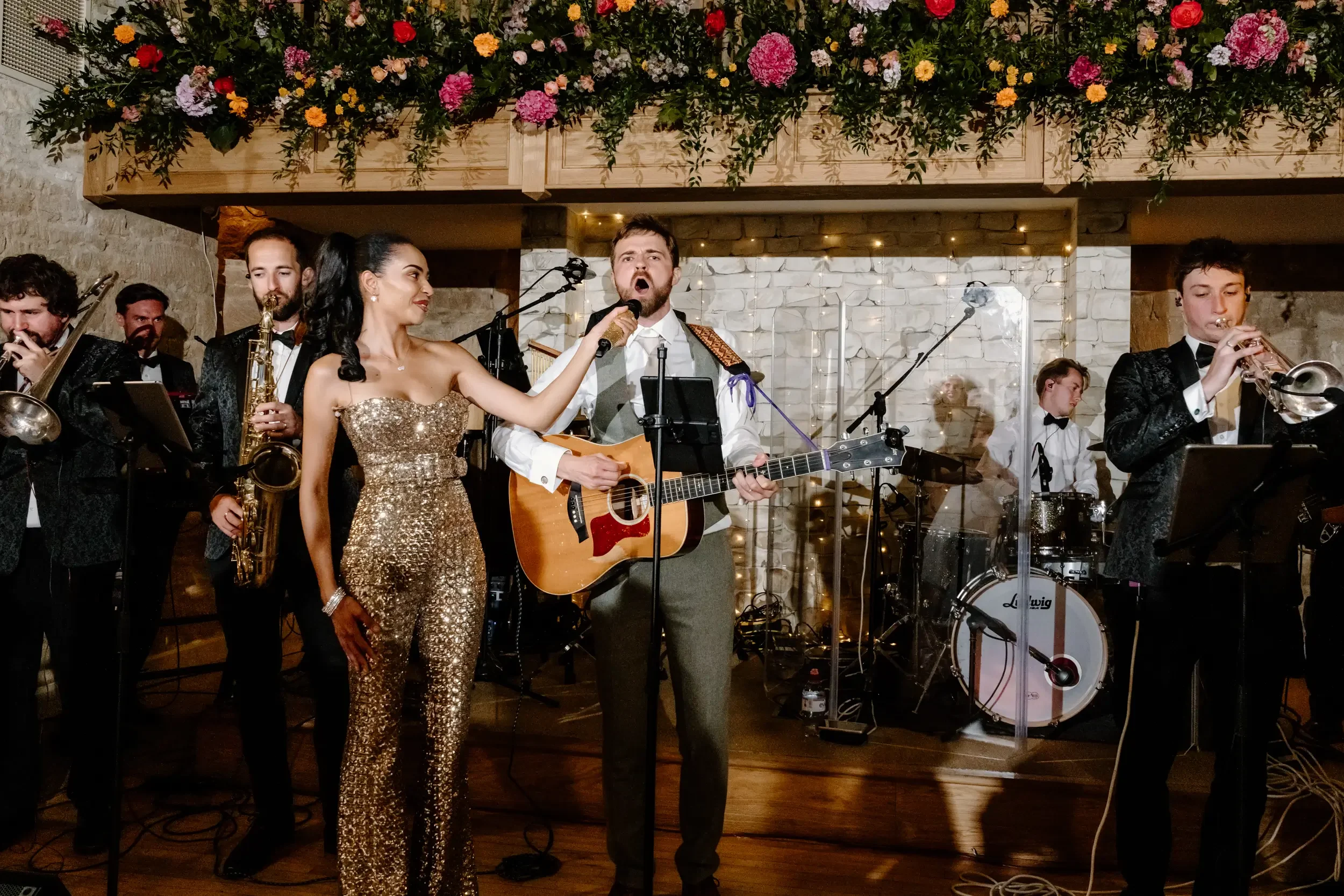 A live band performing on stage at a wedding or celebration, featuring a singer holding a microphone, a guitarist, saxophonist, trumpet player, and drummer, with floral decorations above.