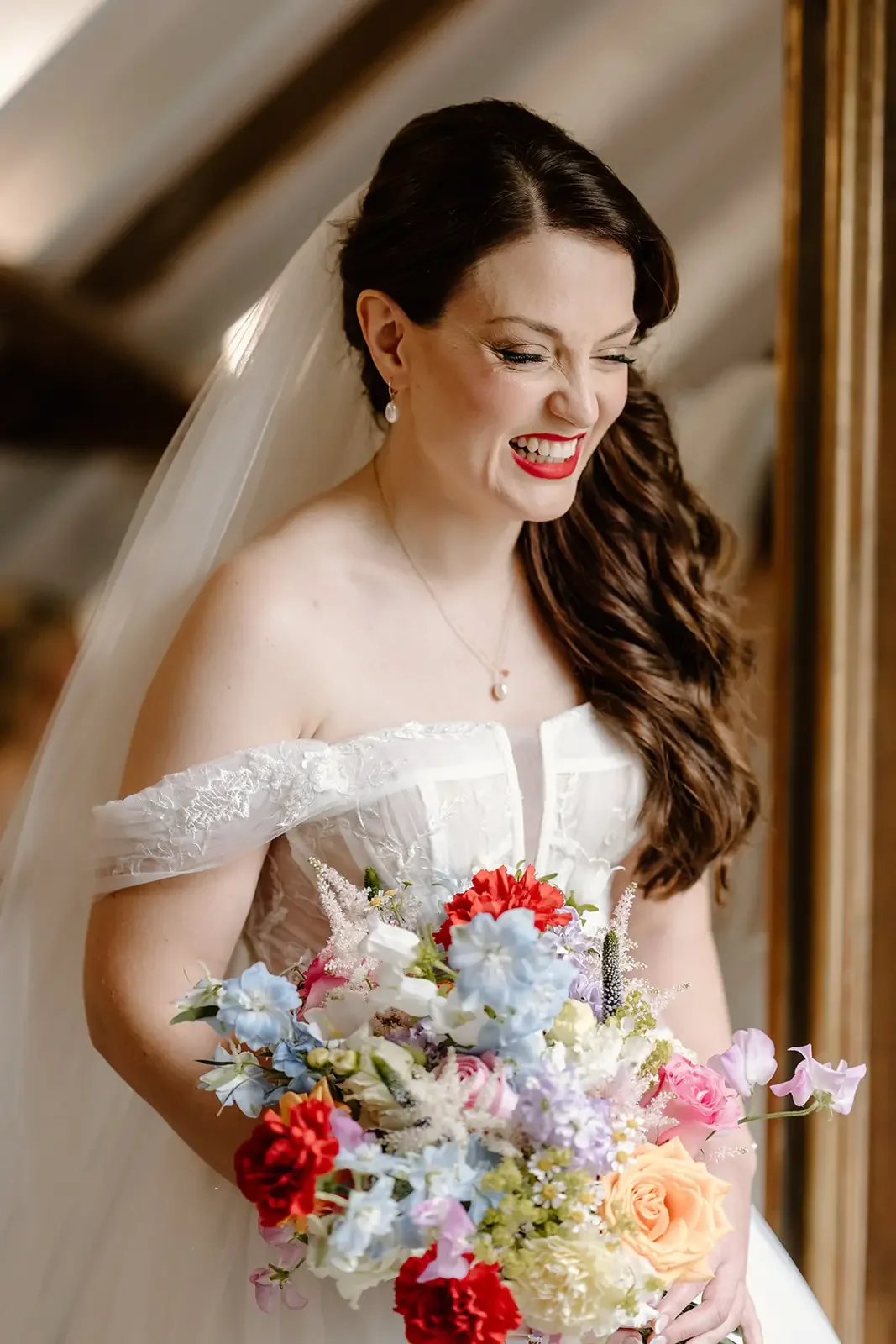 A bride with dark brown hair styled in loose curls, wearing a white wedding gown with off-the-shoulder lace sleeves, holding a colorful bouquet of flowers, and smiling while looking down.