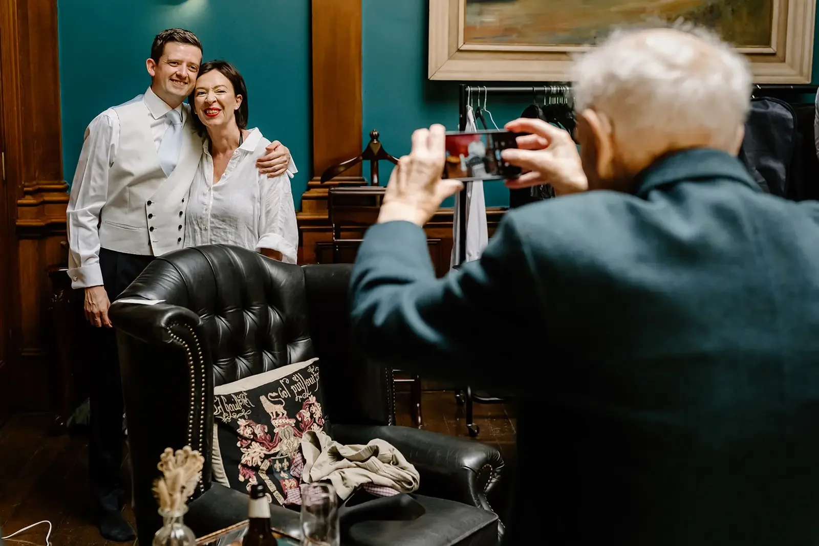 Two people, a man and a woman, are standing and smiling for a photo being taken by an older man with gray hair, in a room with wooden walls and a teal-colored wall. The man has short dark hair and is wearing a white shirt. The woman has shoulder-leng