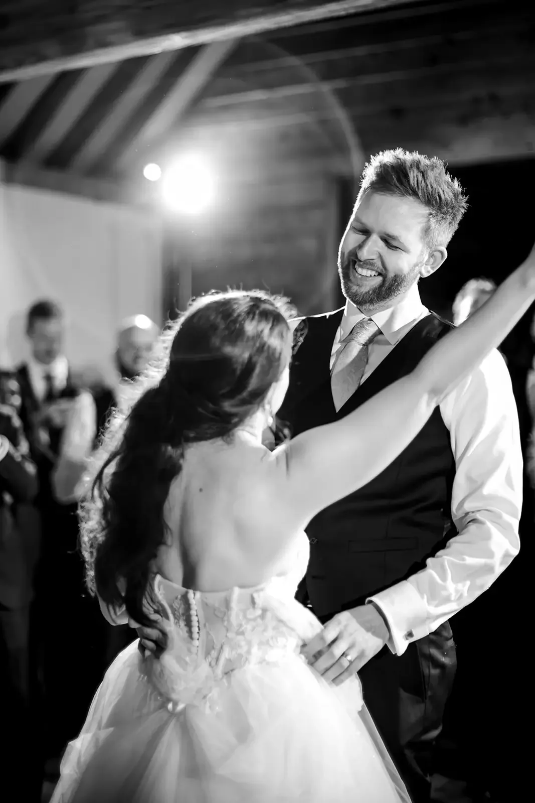 A man and woman in wedding attire dancing at a wedding reception in a barn-like venue with wooden ceiling beams, with guests in the background.