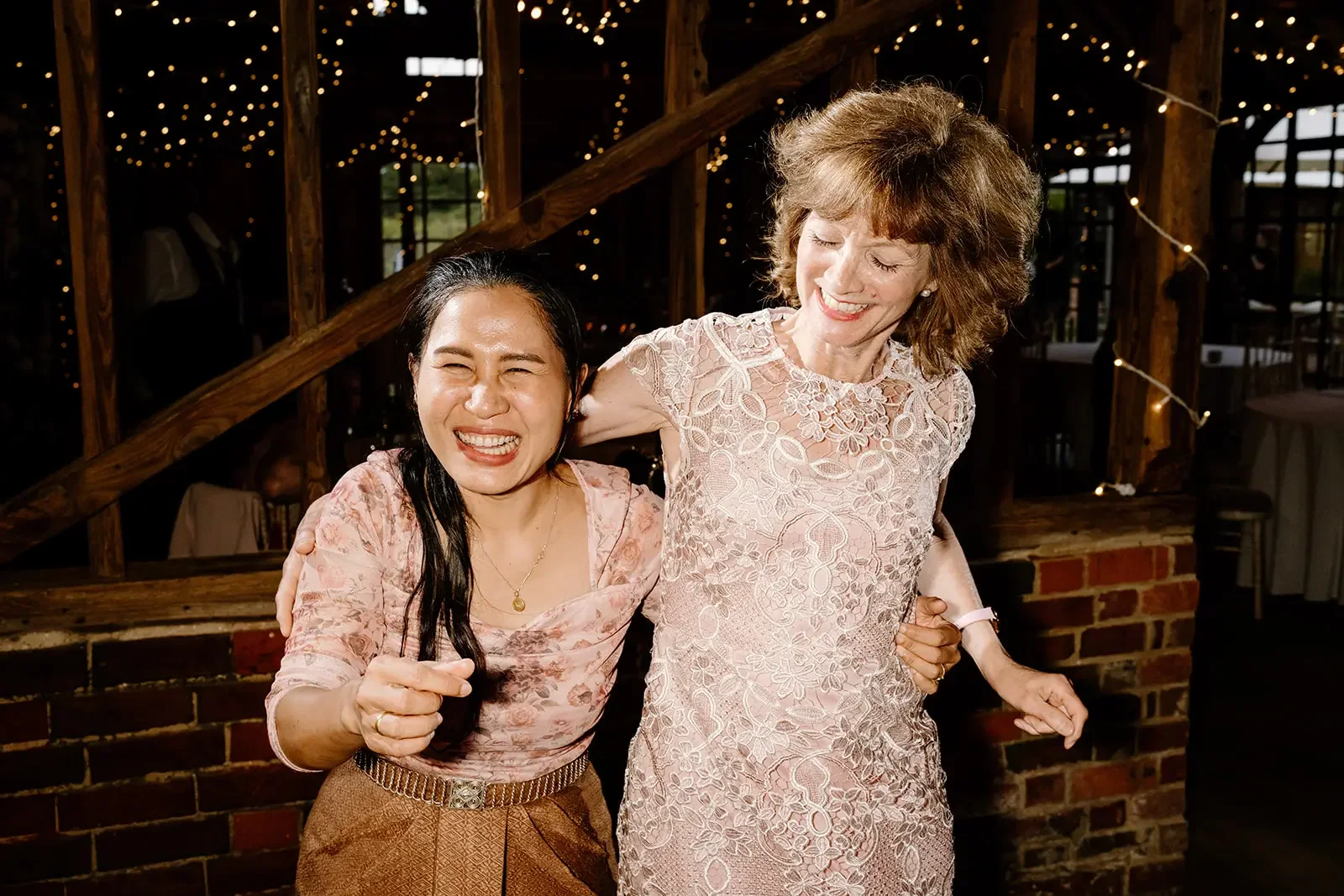 Two women smiling and celebrating at an event, one with dark hair wearing a patterned top and gold belt, the other with red hair wearing a lace dress, standing close together with their arms around each other, in a decorated indoor venue with string 