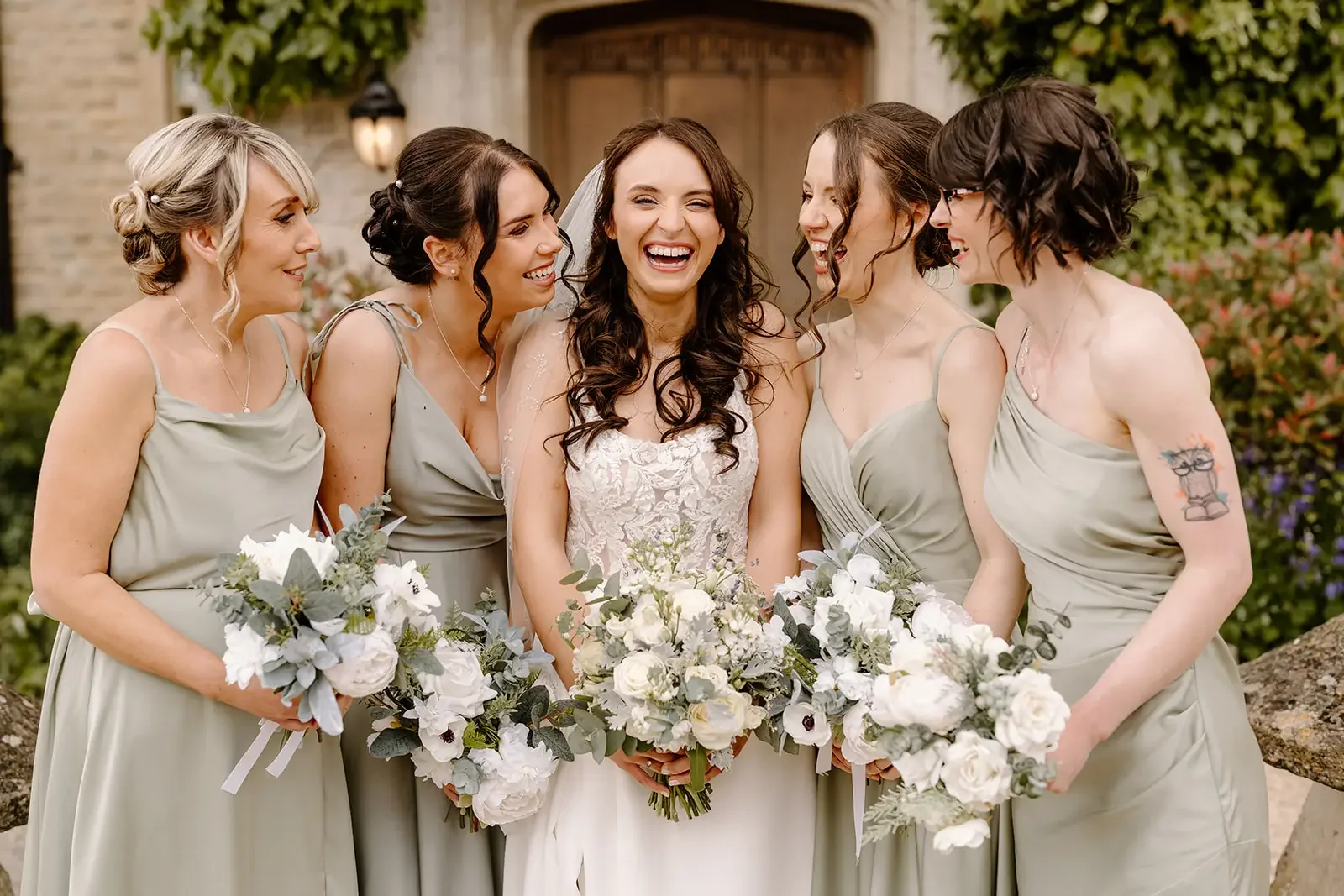 A bride in a white wedding dress surrounded by five bridesmaids in grey dresses, all holding bouquets of white flowers, standing outdoors in front of a building with greenery.