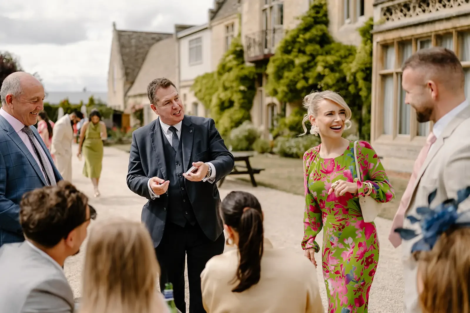 Group of well-dressed people socializing outdoors near a historic building with lush greenery.