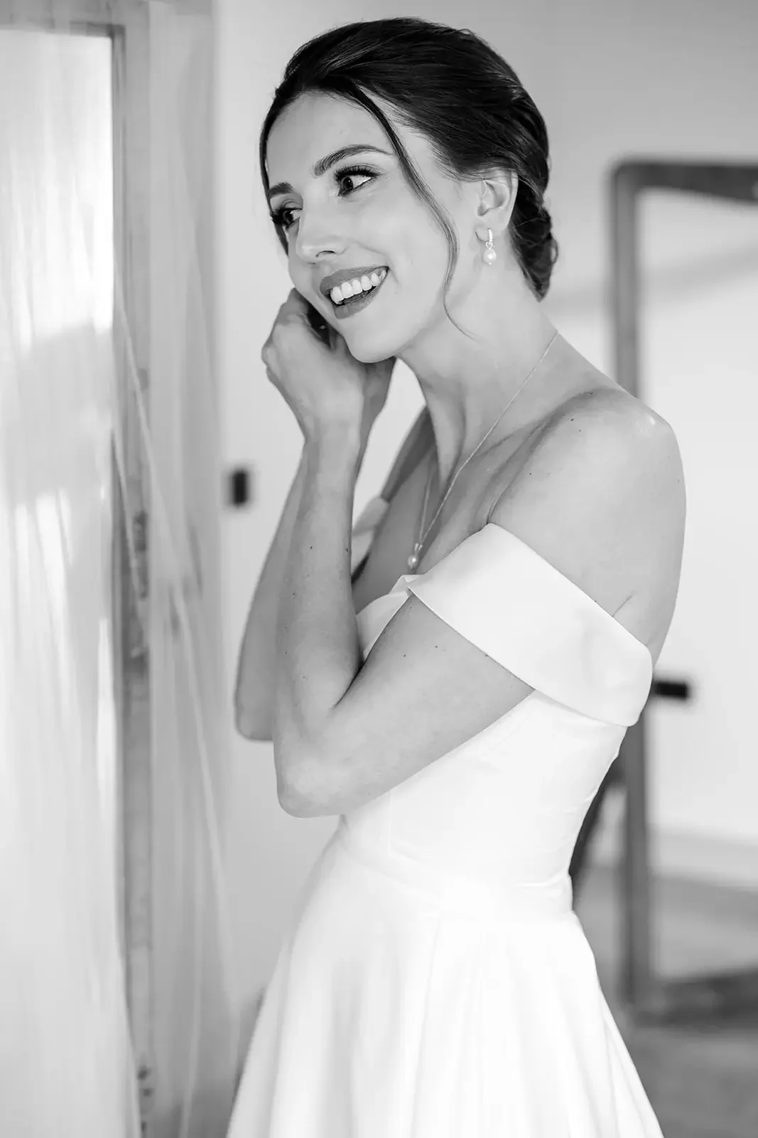 A smiling woman in a wedding dress, with dark hair styled up, standing indoors near a curtain, adjusting her earring.