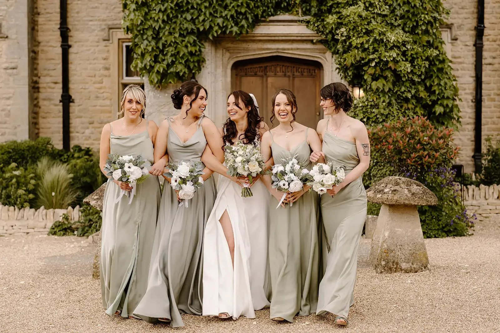 A bride and five bridesmaids, dressed in light grey gowns, standing outside in front of a stone building with greenery, holding bouquets of white flowers, smiling and talking to each other.