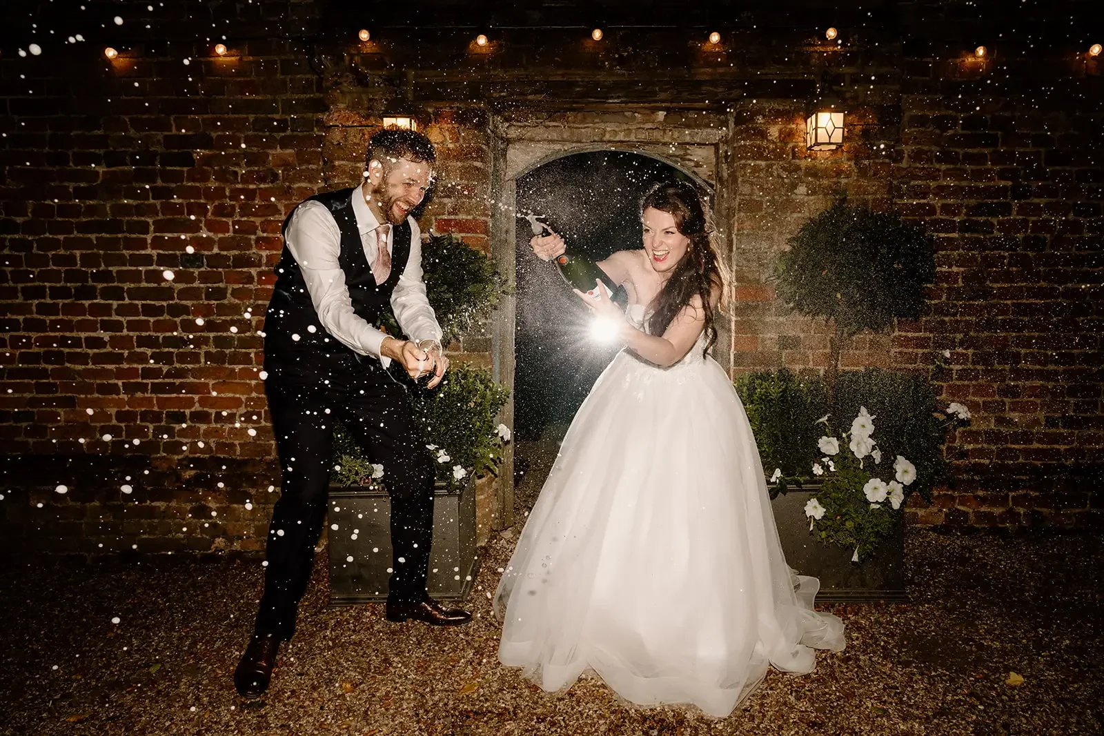 A newlywed couple celebrating their wedding by spraying champagne outdoors at night, with a brick wall and plants in the background.