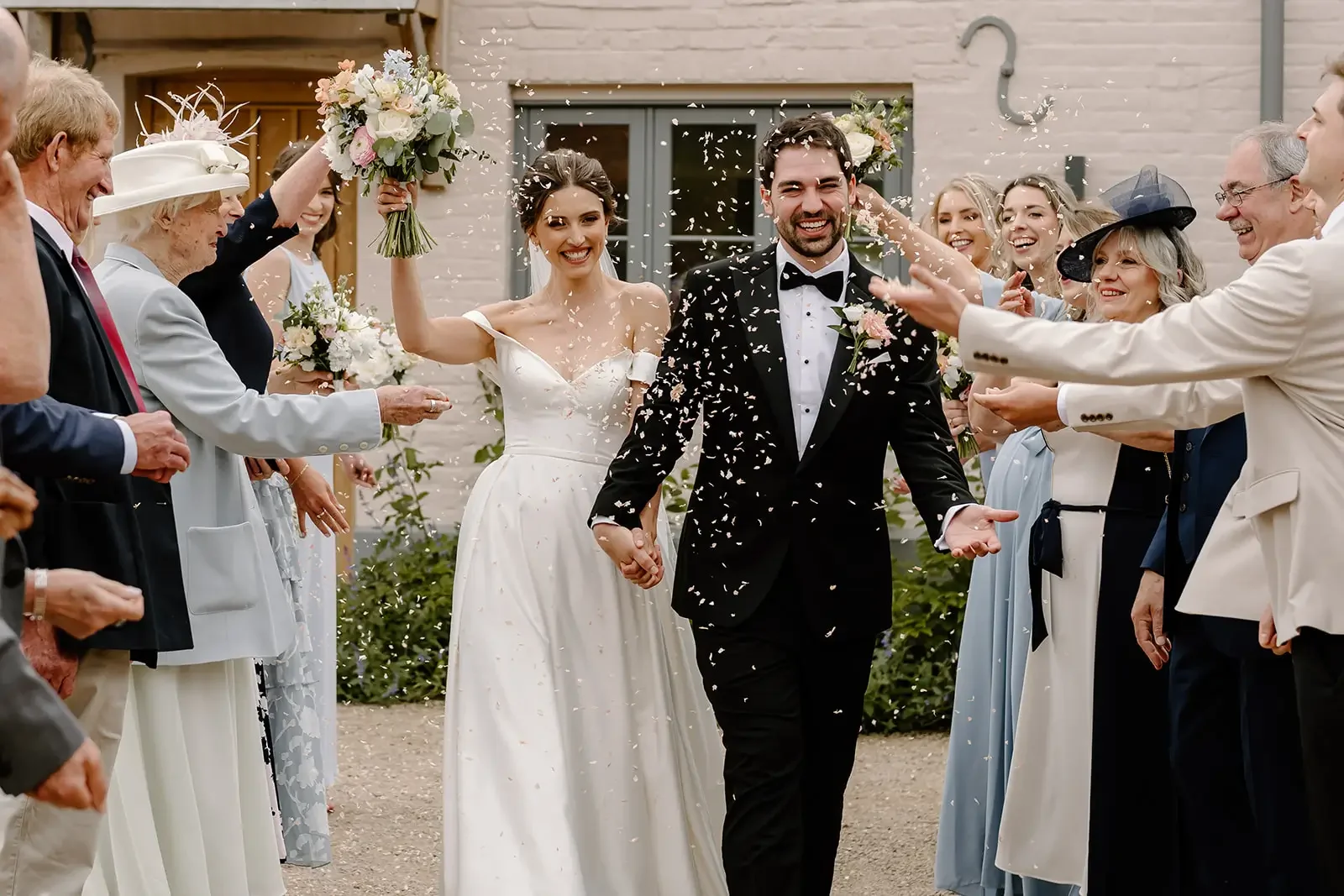 A newlywed couple celebrates their wedding with friends and family outside, surrounded by confetti. The bride is holding a bouquet and wearing a white wedding dress, while the groom is in a black tuxedo. Guests are joyfully throwing confetti and smil