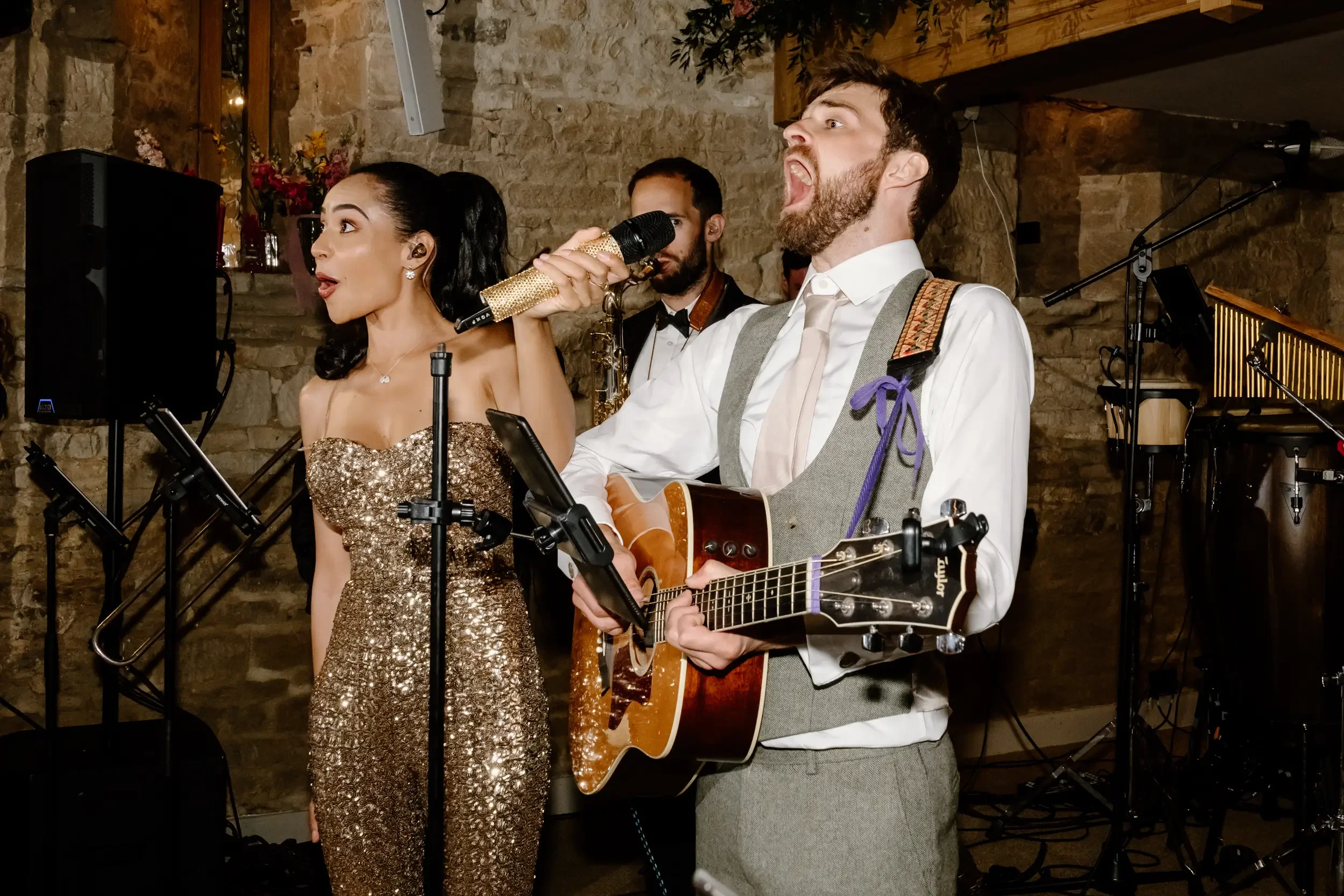 A man with a beard, wearing a white shirt and a gray vest, is singing into a microphone and playing an acoustic guitar. A woman in a gold, glittery strapless dress is standing next to him, holding a microphone and singing. In the background, there is
