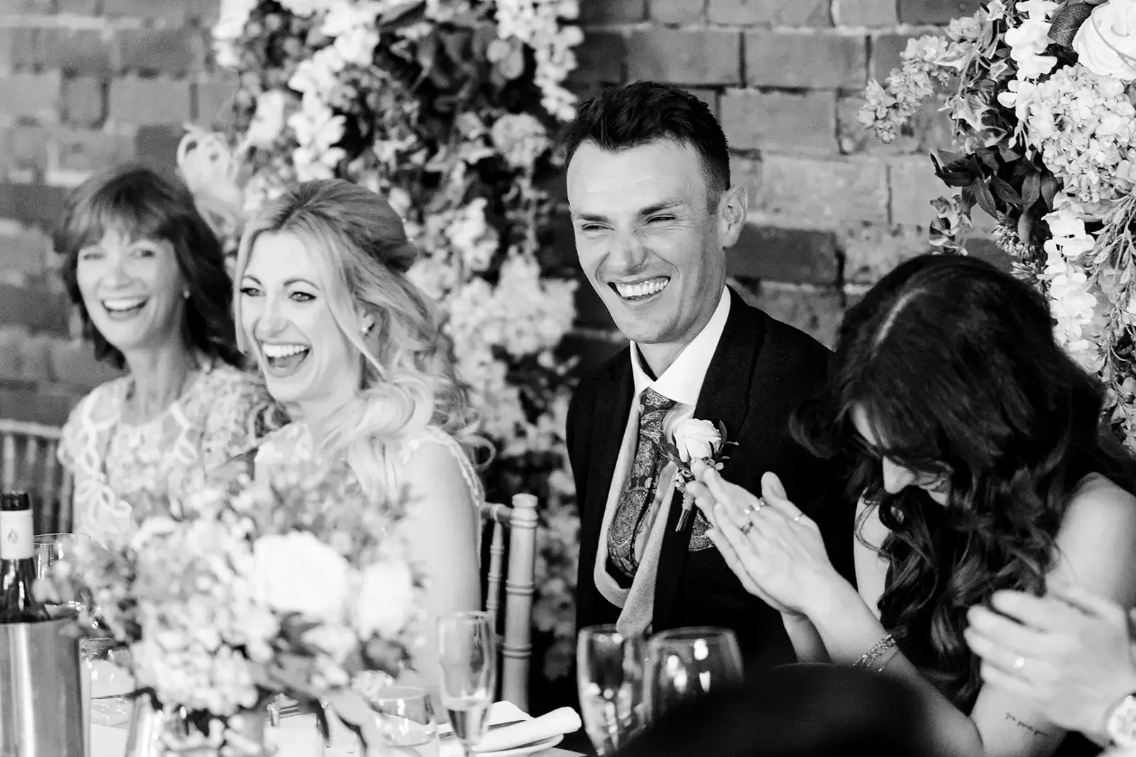 People at a wedding reception sitting at a table decorated with flowers; one man in a tuxedo is laughing, two women beside him are also laughing, and another woman is clapping her hands.