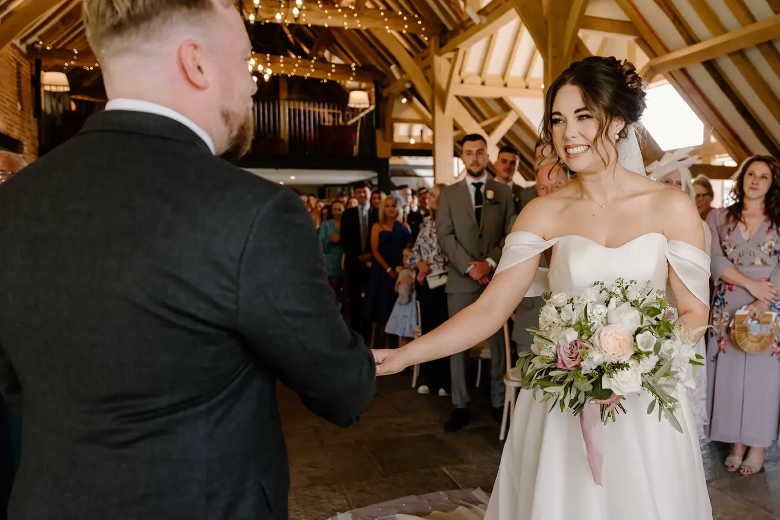 A bride and groom during their wedding ceremony, holding hands and smiling at each other inside a wooden, rustic venue with guests in the background.
