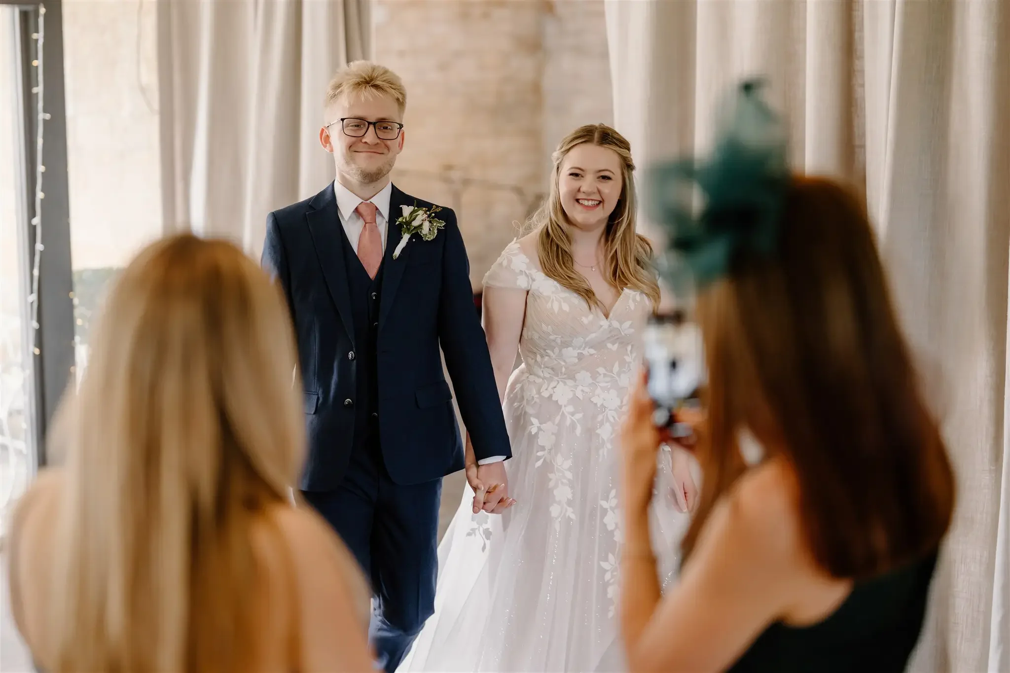 Bride and groom holding hands during their wedding ceremony, smiling, with two women taking photos in front of them.